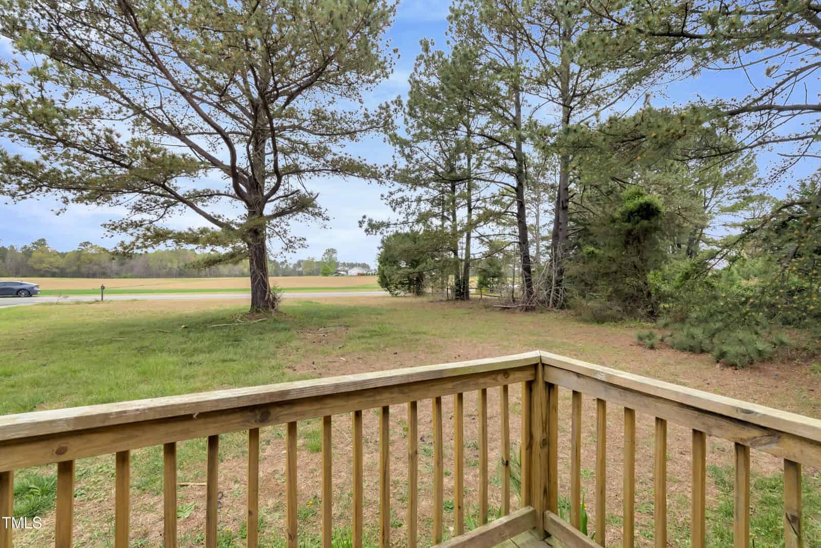 View from a wooden deck overlooking a grassy yard with several tall pine trees. A car is parked on a road to the left. The sky is partly cloudy, and the scene conveys a peaceful rural setting.