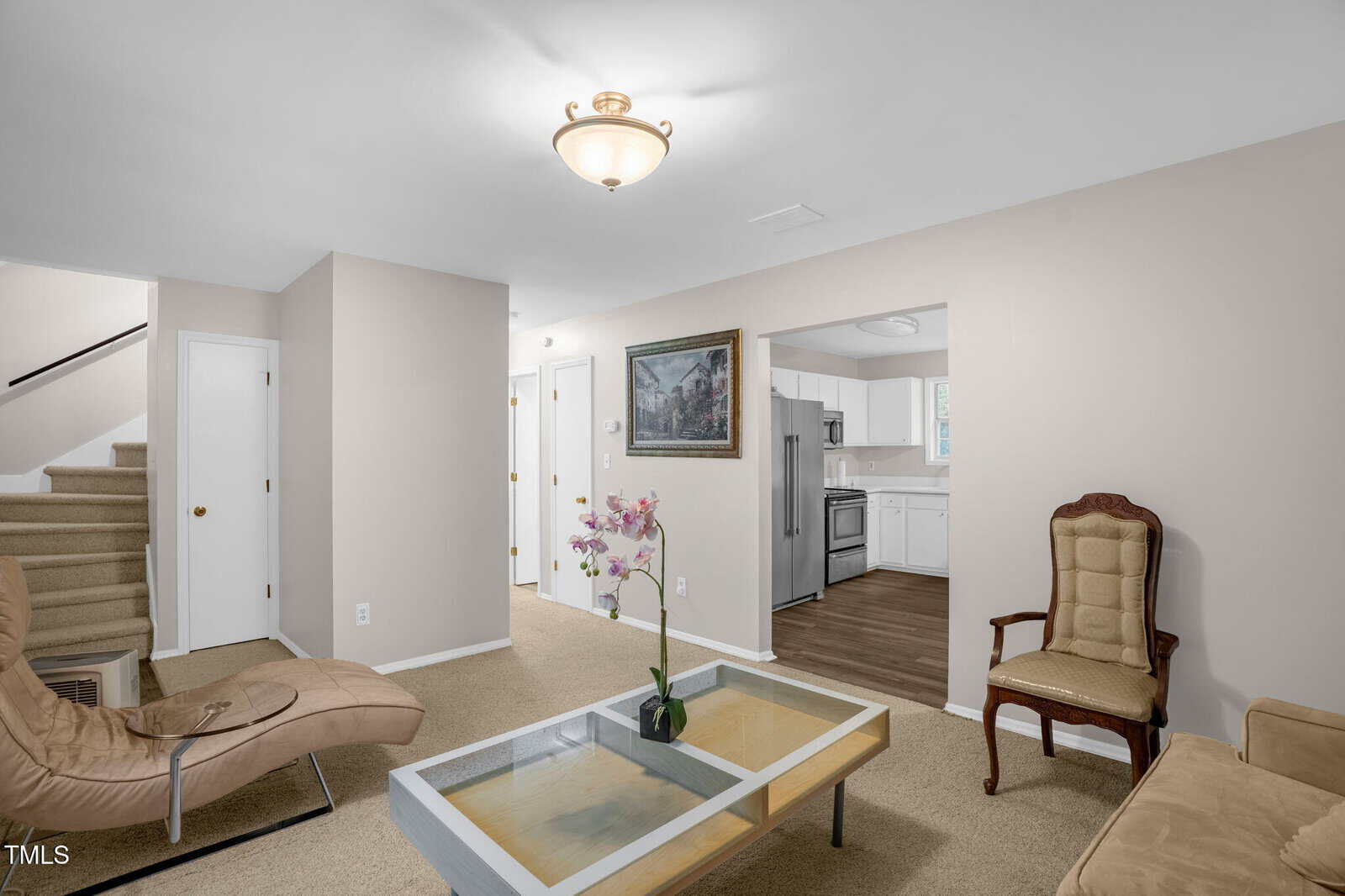 A living room with beige walls and carpet, featuring a glass coffee table with a pink flower arrangement. There's a cushioned chair, a modern recliner, and a view into a kitchen with white cabinets and wooden flooring. A staircase is visible to the left.