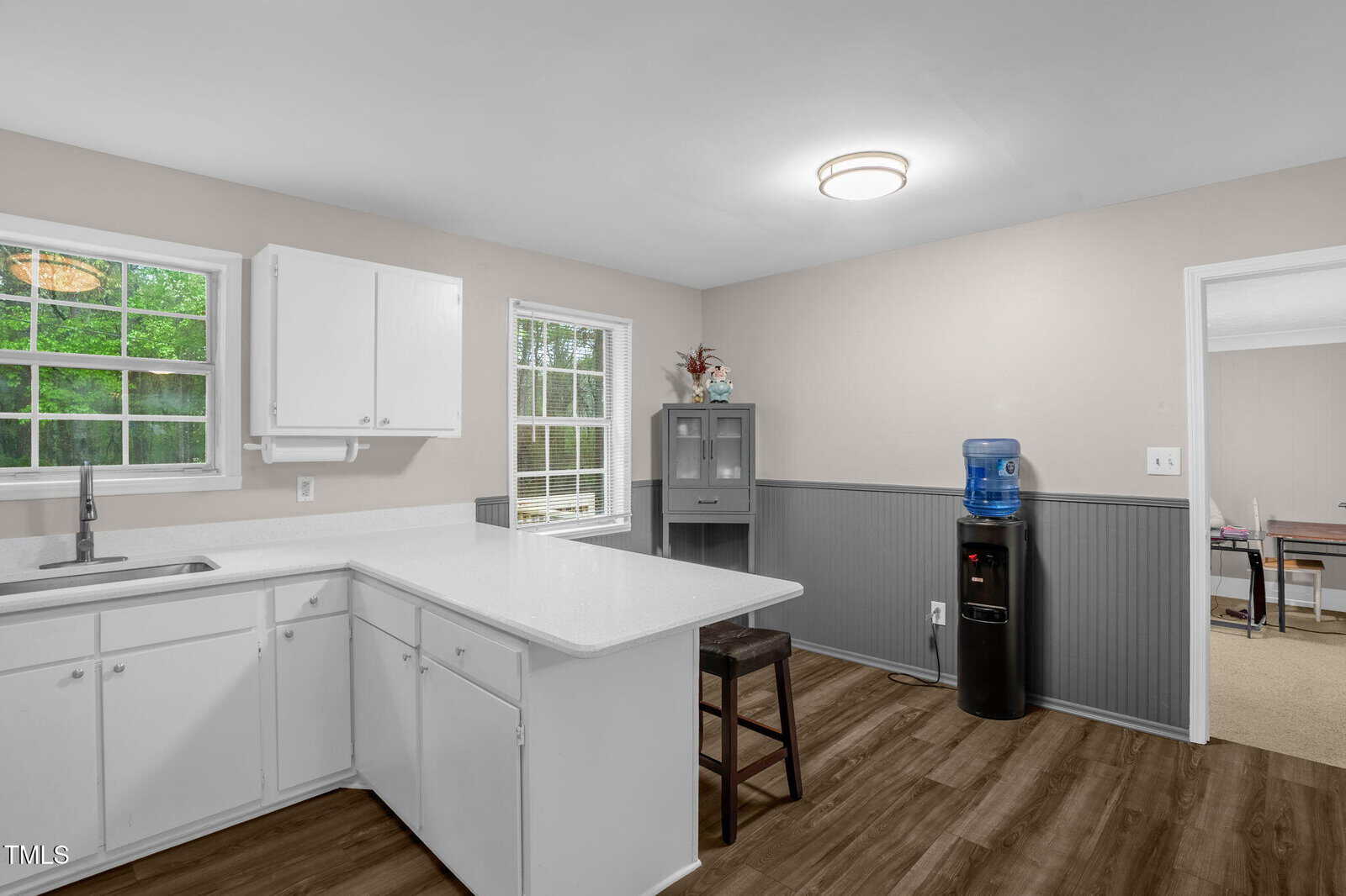 A kitchen with white cabinets and countertops, featuring a sink, under a ceiling light. There's a window above the counter. A water cooler stands to the right, near a small cabinet, with an entrance to another room visible in the background.