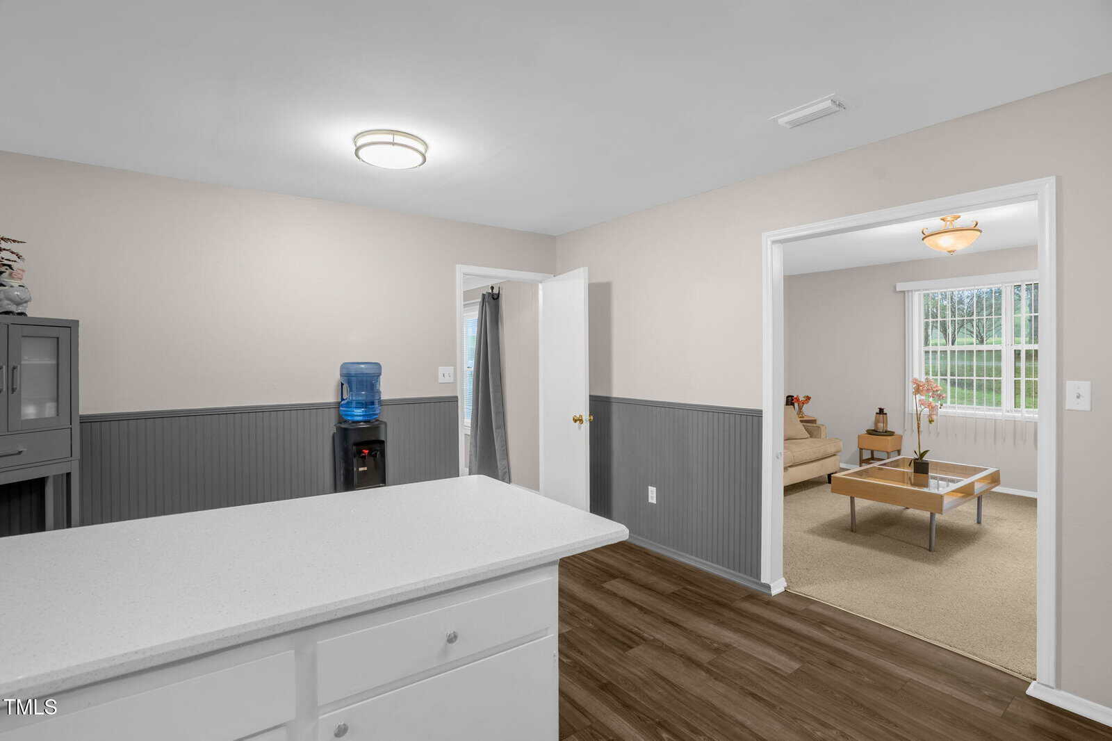 Kitchen with white cabinets and a countertop, leading to a living room with a beige sofa and wood table. Walls are adorned with wainscoting, and a water cooler is near the door. A light fixture is on the ceiling, and the flooring is wood-style.