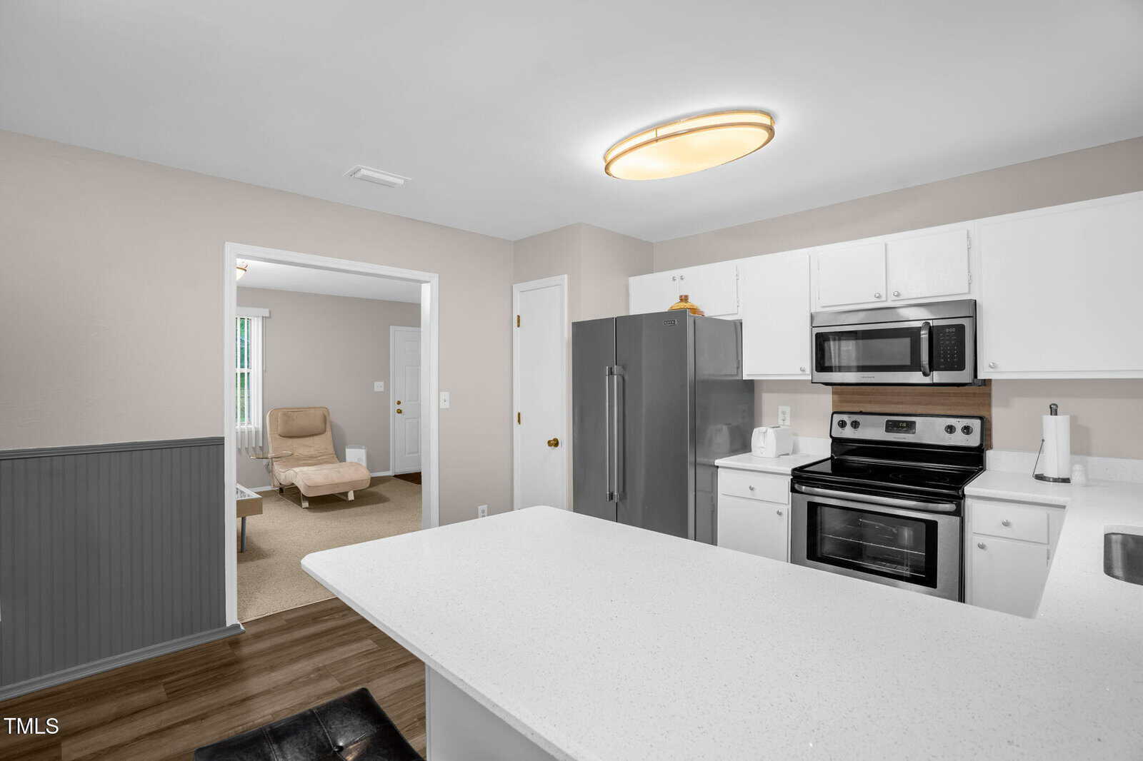 A kitchen with white cabinets, a silver refrigerator, and a stainless steel oven and microwave. A large white countertop is in the foreground. An adjoining room with beige walls and carpet is visible, furnished with a beige chair and a coffee table.