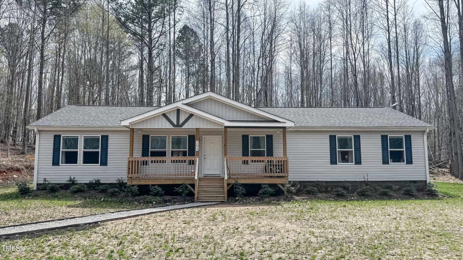Nestled on Oxford's 3014 Split Wood Court, this charming one-story house boasts a light gray exterior, dark shutters, and a quaint wooden front porch. Tall, leafless trees surround the home, set on a grassy lot with a narrow gravel path leading to the entrance.