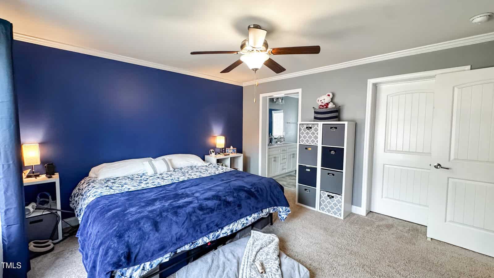 A bedroom featuring a bed with blue bedding against a dark blue accent wall. A ceiling fan with lights is above. A white door and a storage unit with cubbies to the right. An open door leads to a bathroom. Carpeted floor with an open suitcase.