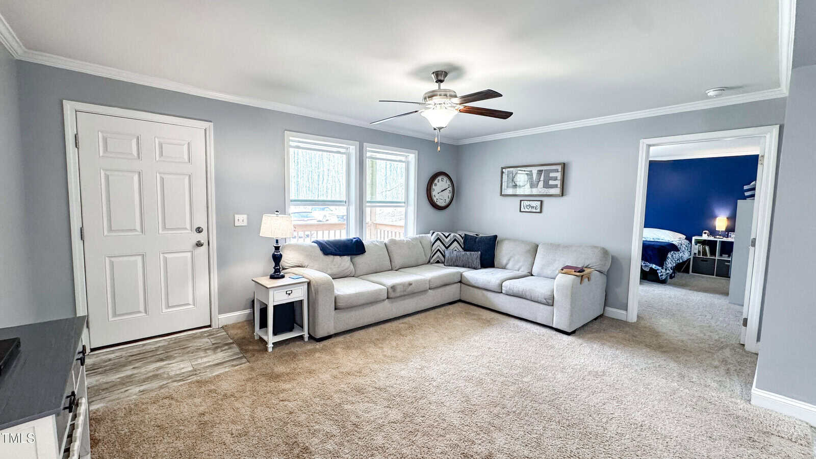 A living room with gray walls and carpet flooring. It features a large gray sectional sofa, a small side table with a lamp, a ceiling fan, and a wall clock. A doorway leads to another room with a blue accent wall.