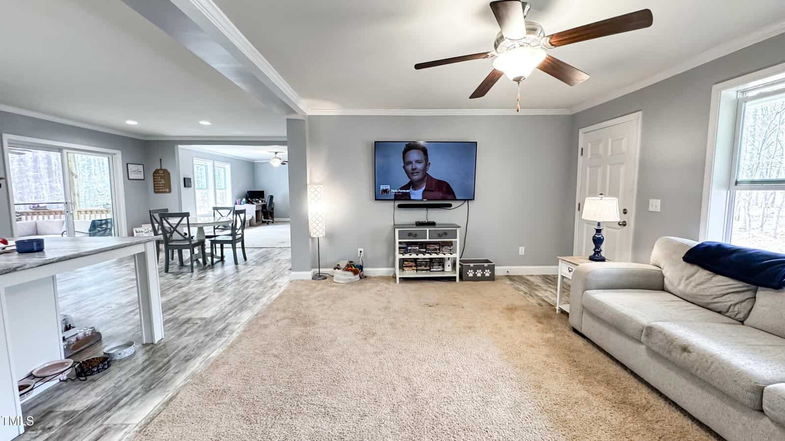 Living room with beige carpet and gray walls, featuring a wall-mounted TV, white ceiling fan, and a light gray sofa. A small white side table with a lamp is next to the sofa. The room connects to an open dining area and has large windows.