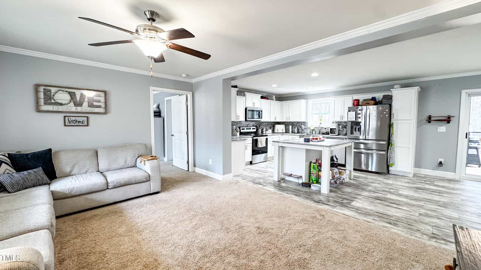 Open-concept living space with gray walls, featuring a white kitchen with an island, stainless steel appliances, and wooden flooring. The adjacent carpeted living area has a beige sectional sofa and a ceiling fan. Wall decor includes a "LOVE" sign.
