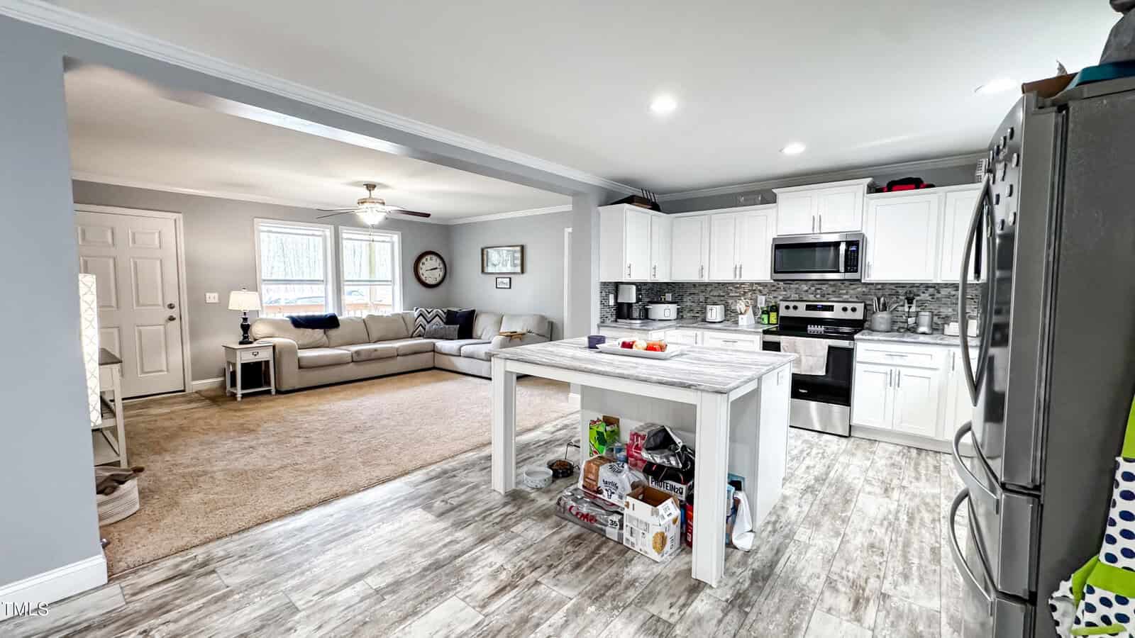 A kitchen with white cabinetry, stainless steel appliances, and a marble island opens into a living room with a beige sofa and wall clock. The floor is wood, and the counter has fruit and various items. Bright windows illuminate the space.