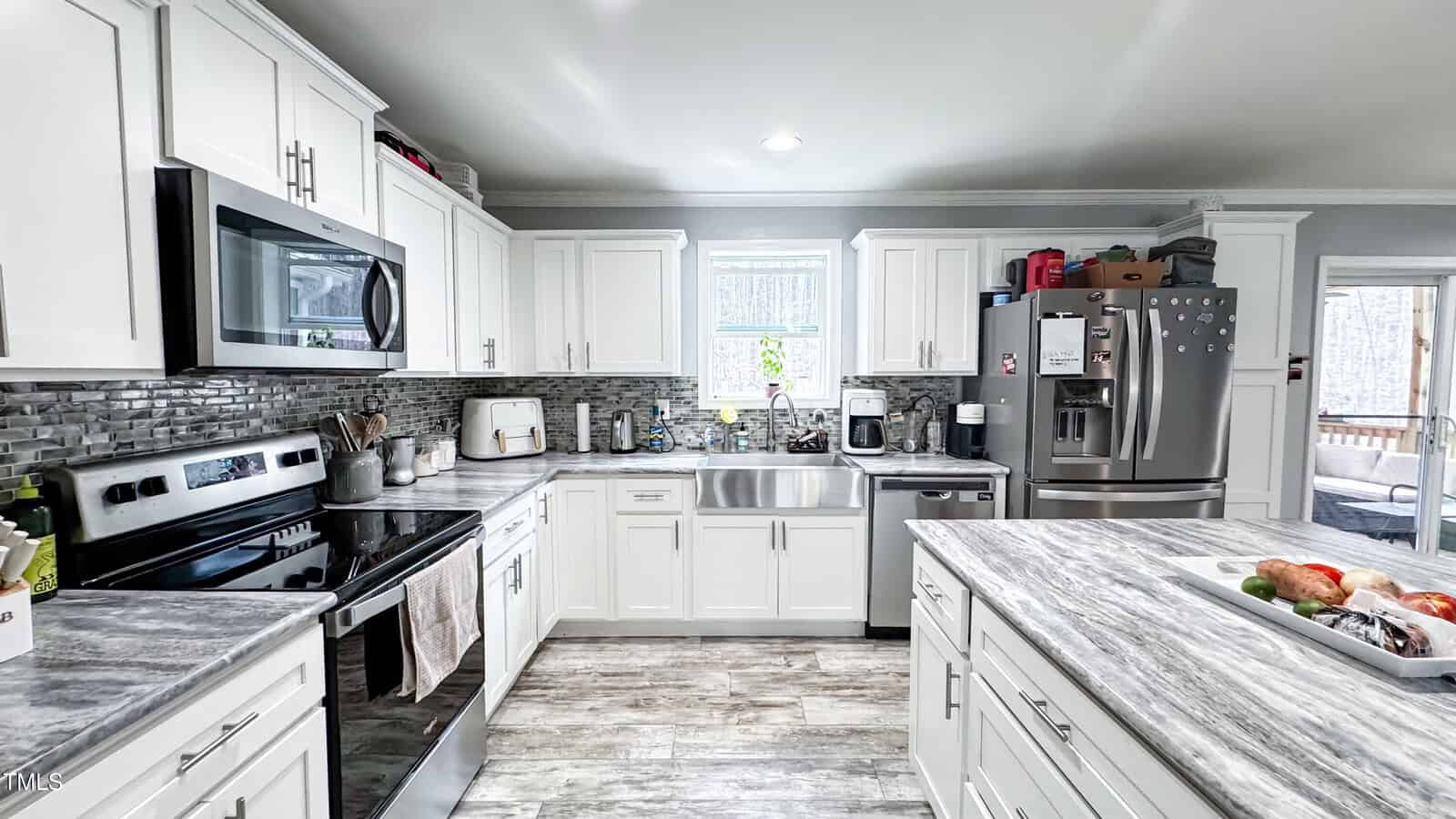 A modern kitchen with white cabinets and gray stone countertops. Stainless steel appliances, including a refrigerator, microwave, and oven, are present. A small window is above the sink. The flooring is wood, and various kitchen items are on the counters.