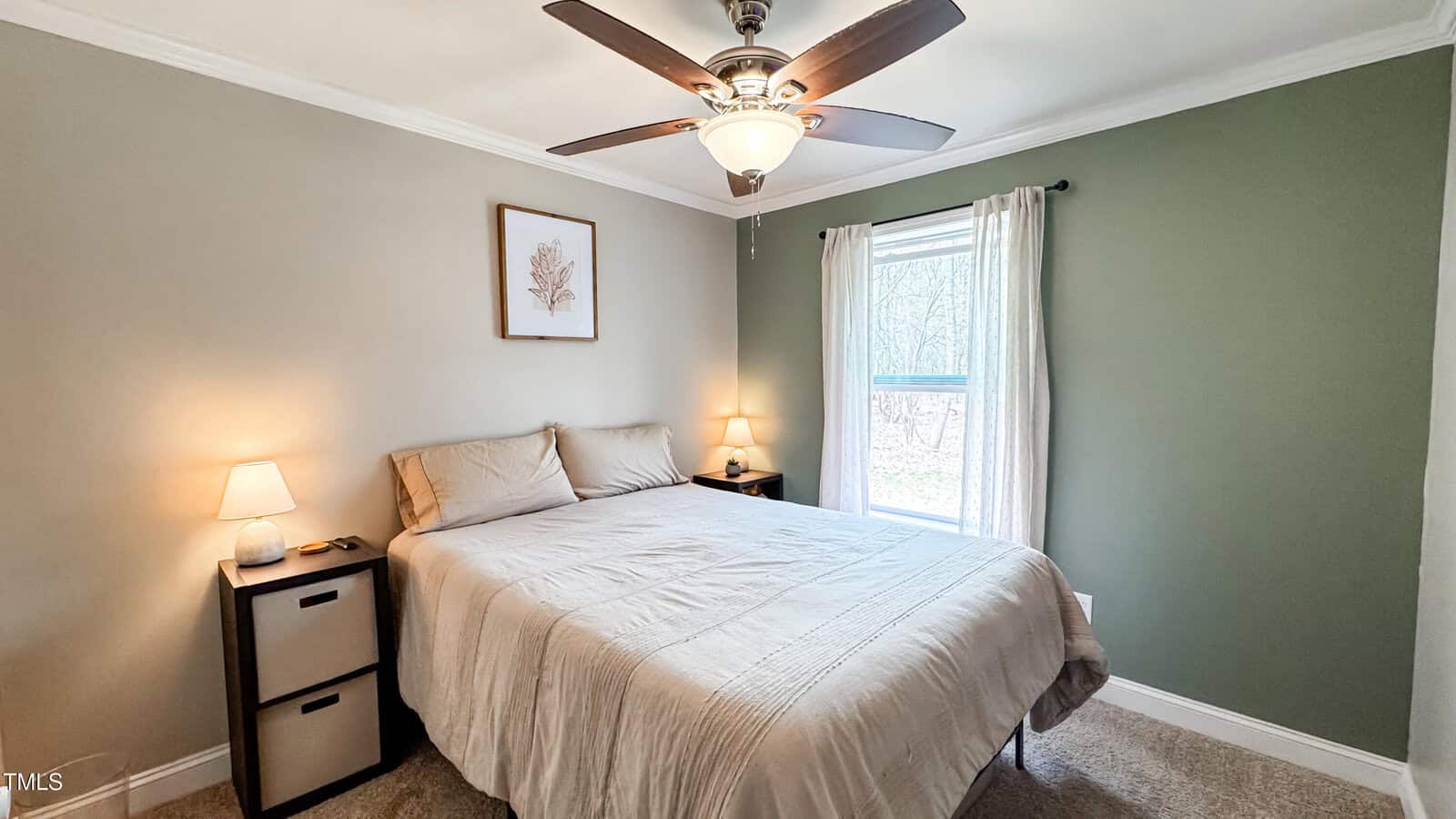 A bedroom features a large bed with beige bedding, framed leaf artwork above, and two small bedside tables with lamps. A ceiling fan is mounted above, and a window with a light curtain allows natural light into the room with green and beige walls.