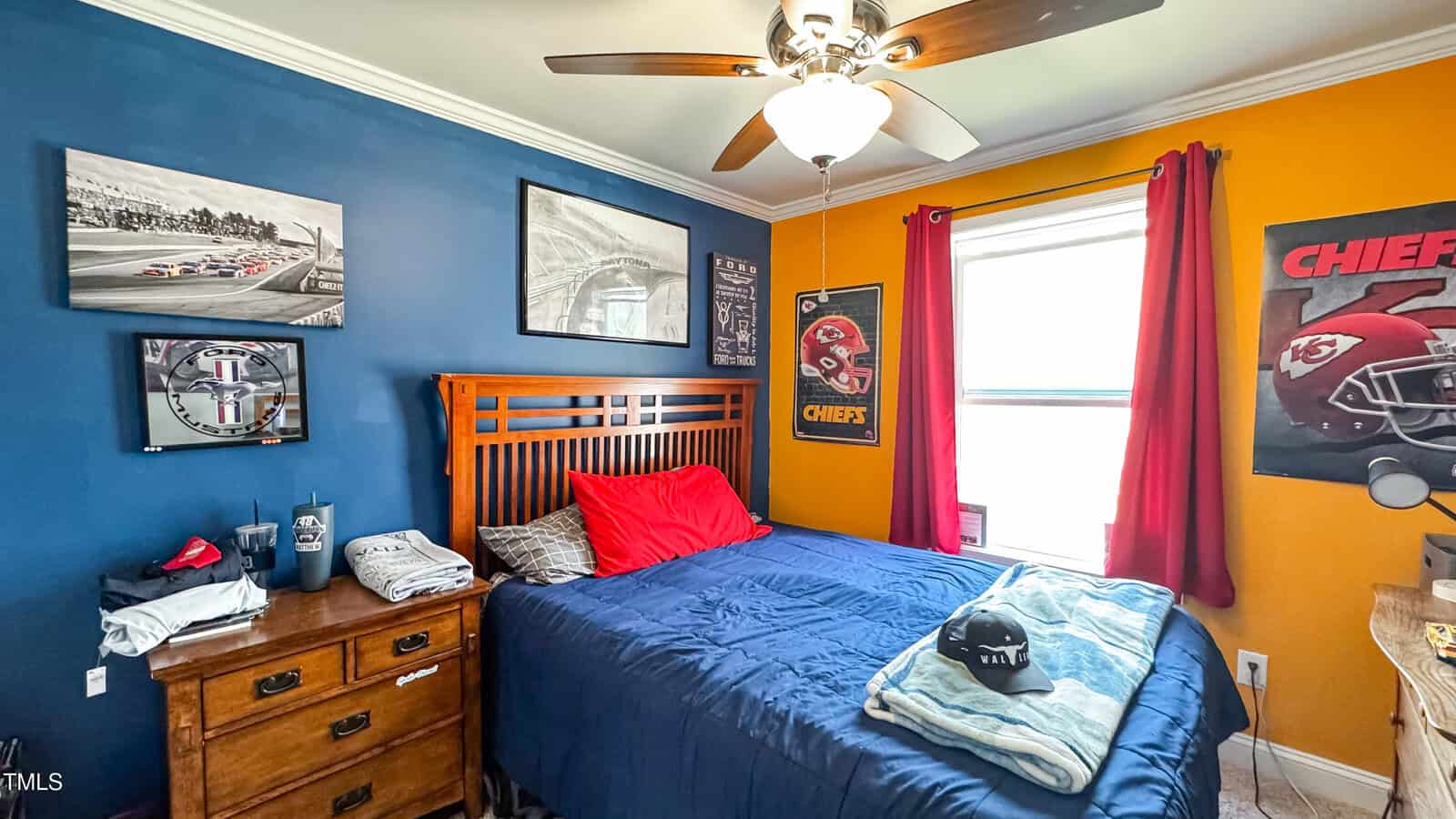 A bedroom with a wooden bed covered in blue bedding and a red pillow. The walls are blue and yellow, adorned with sports posters and framed images. A ceiling fan with a light hangs above, and red curtains frame a window. A dresser is on the left.