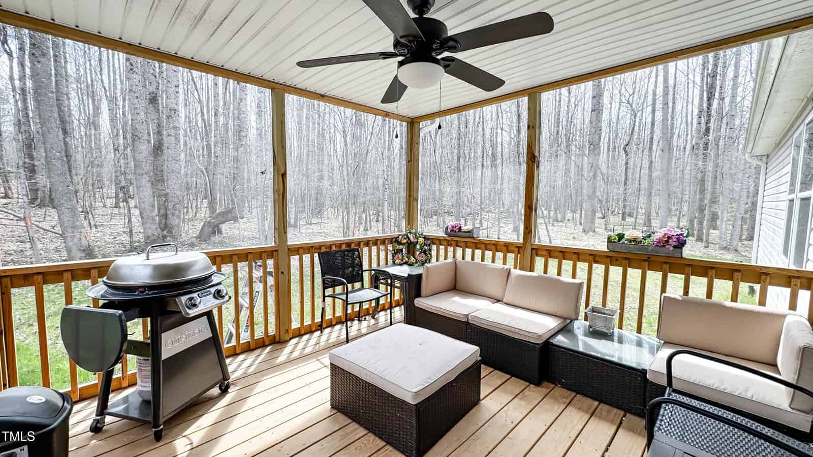 Screened porch with wicker furniture, including a sectional sofa and ottoman with light cushions. A grill is on the left. There is a ceiling fan above, and the background shows a view of leafless trees. Potted plants are on the railing.