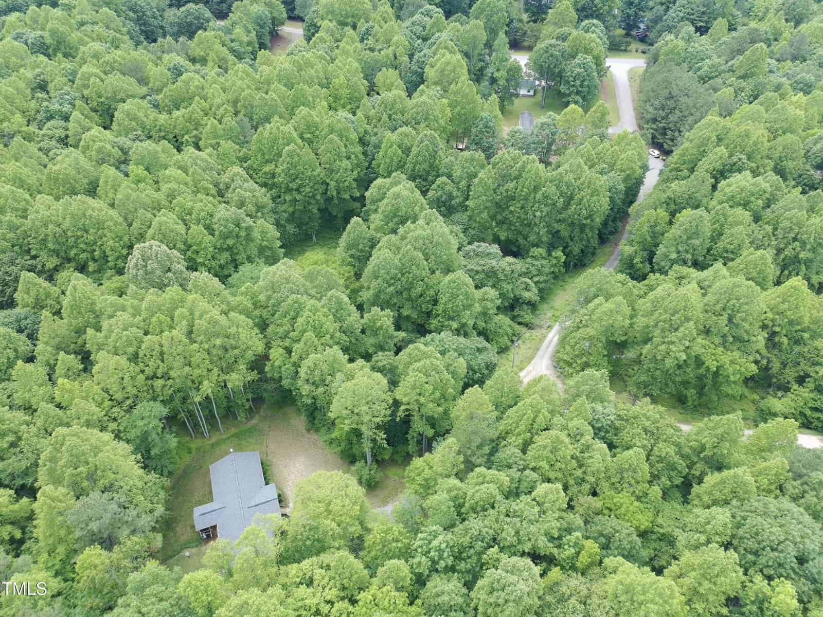 Aerial view of a house with a grey roof surrounded by dense green trees, with winding roads visible among the forested area. Several clearings and structures are also partially visible through the trees.