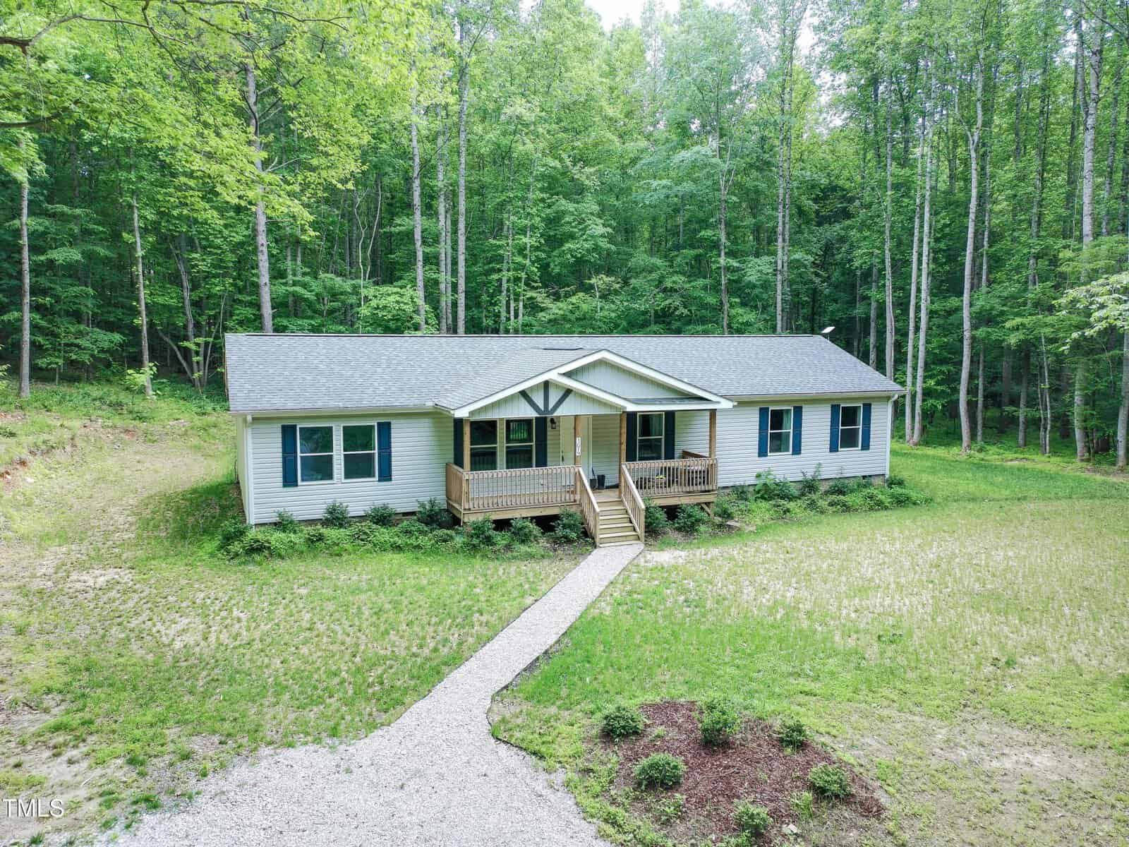 Nestled in a clearing at 3014 Split Wood Court, Oxford, this single-story house features gray siding and a covered front porch. A gravel path winds through tall trees to the front steps, with lawn and small shrubs along the way.