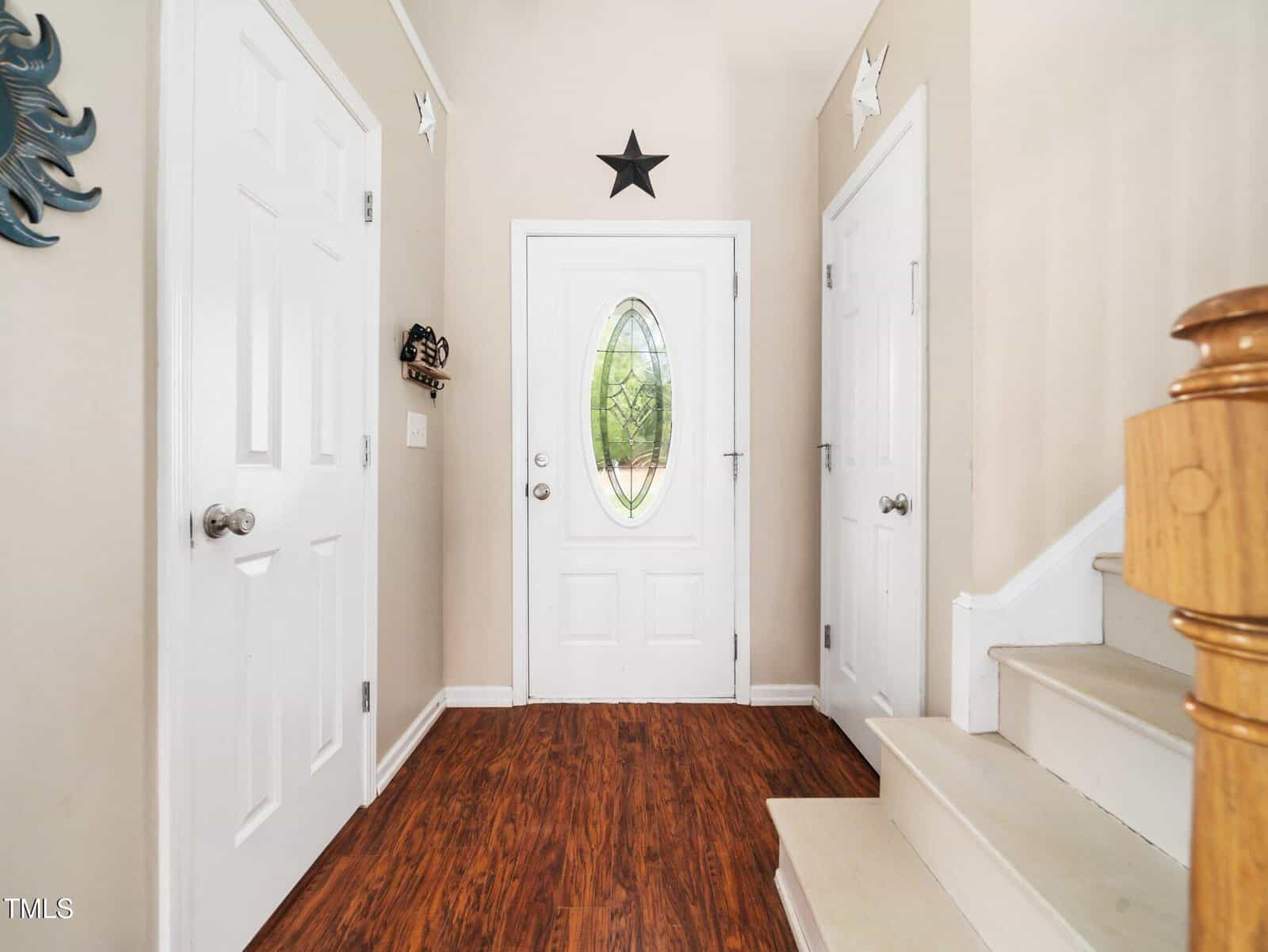 A hallway with a wooden floor features a white front door with an oval glass panel. The walls are light-colored, with two additional white doors on either side. A black star decoration is above the front door. A wooden staircase is on the right.