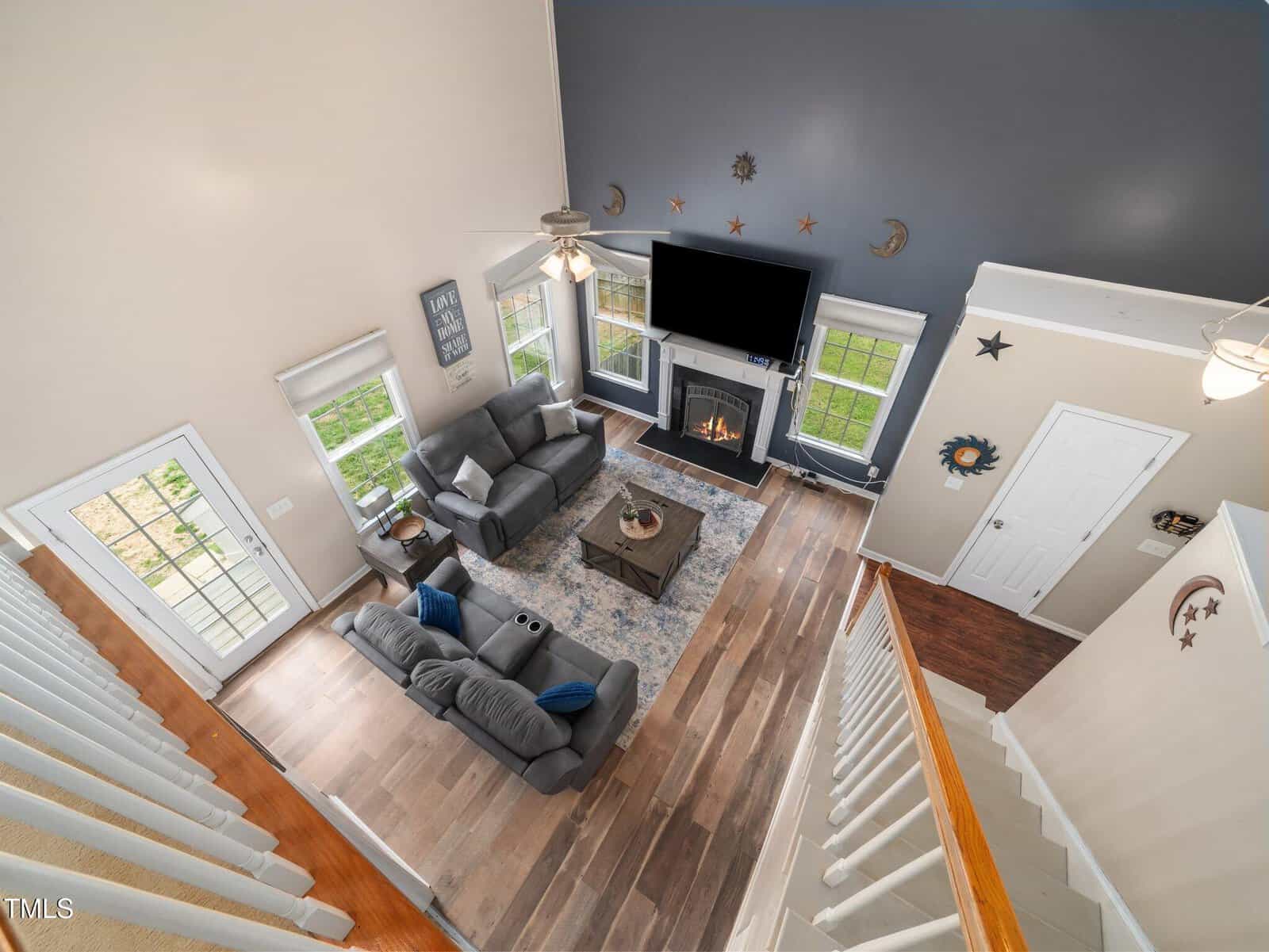 A spacious living room with a high ceiling, viewed from above. It features two gray couches, a coffee table, and a fireplace with a TV above it. The room has wooden flooring, a large rug, and several windows, allowing natural light to enter.