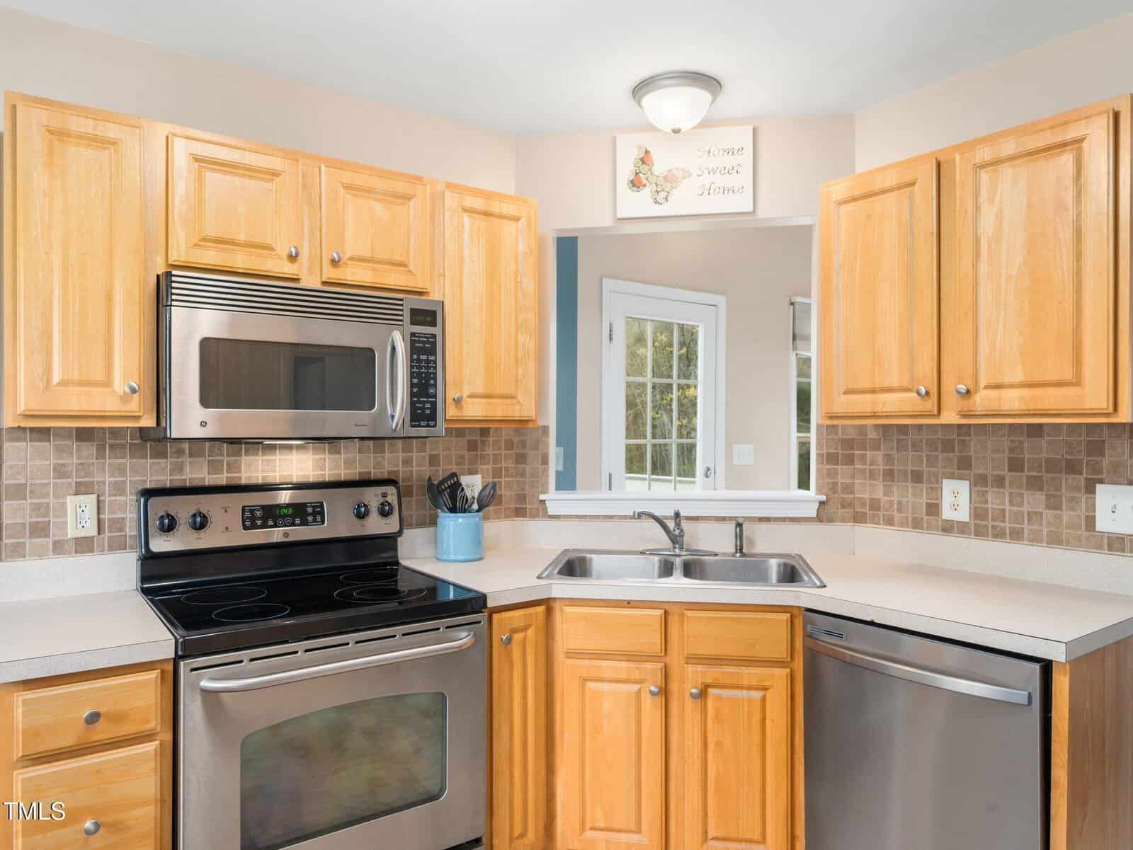 Kitchen with light wood cabinets and beige countertops. Features a stainless steel stove, microwave, and dishwasher. A double sink is below a small window. Beige tile backsplash. A small decorative sign above the sink reads "Home Sweet Home.
