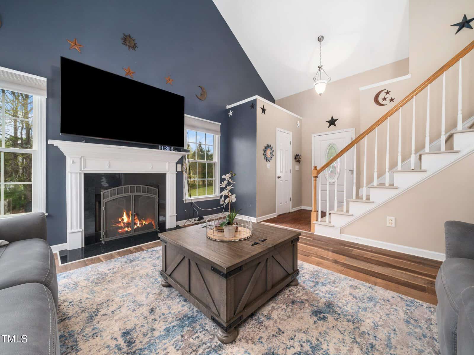 A living room with a vaulted ceiling features a fireplace under a wall-mounted TV. There's a wooden coffee table with a plant, surrounded by gray sofas. The walls have star and moon decorations, and a staircase leads upstairs by the entrance door.