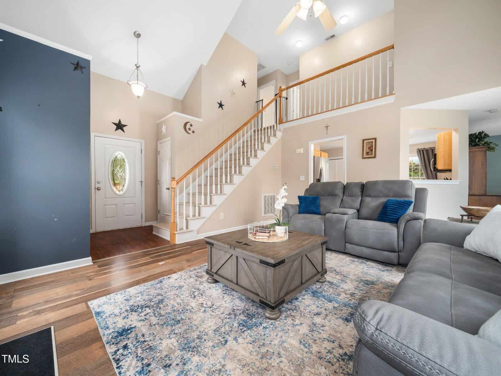 A spacious living room featuring a staircase with white railings and wood steps. It has a gray couch, a patterned rug, and a wooden coffee table with decorative items. A ceiling fan and chandelier hang above. There are star decorations on the walls.