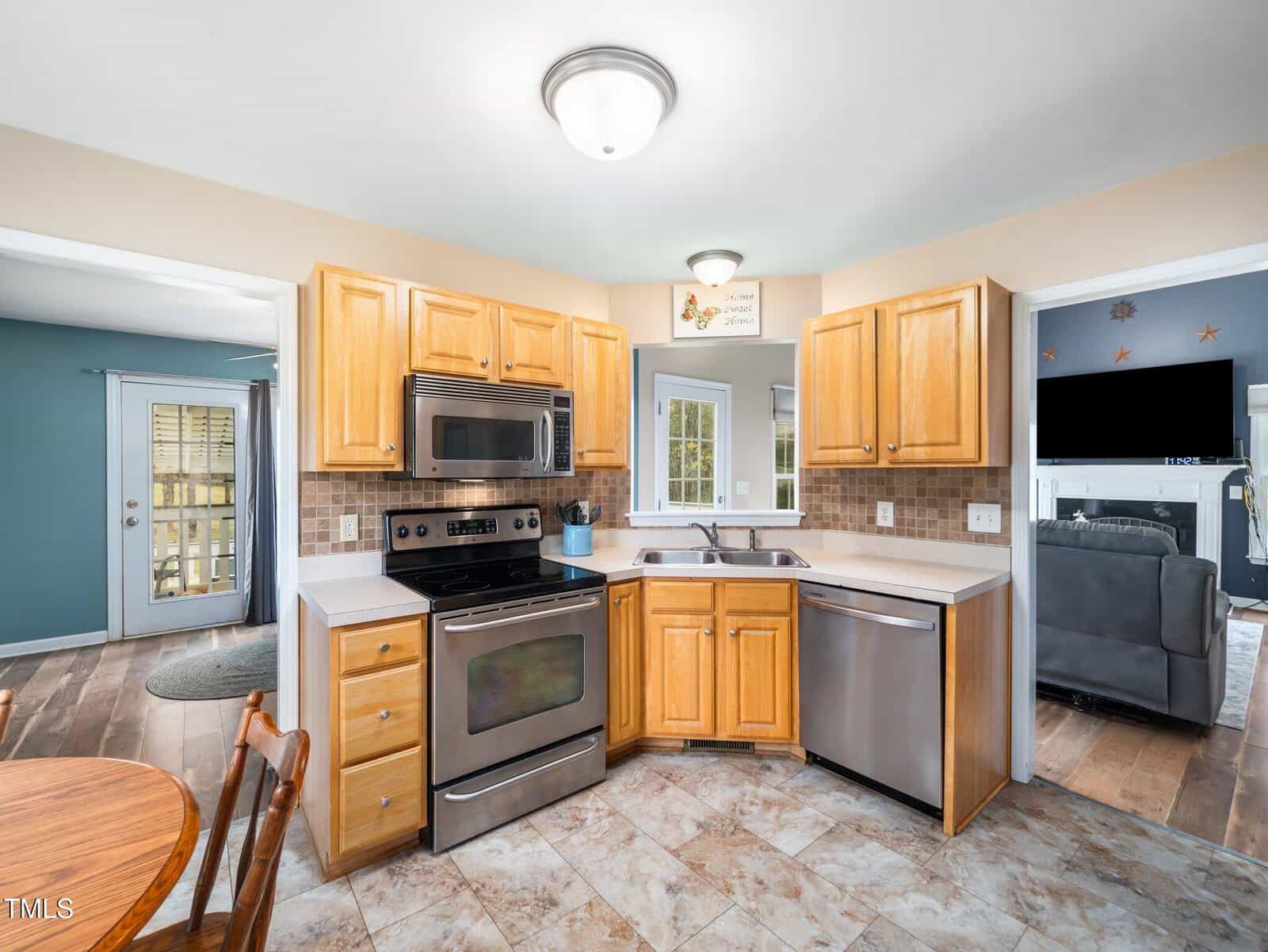 A kitchen with light wood cabinets, stainless steel appliances, and a double sink. There is a stove with an overhead microwave and a dishwasher. The floor is tiled, and two doorways lead to adjacent rooms with wooden flooring.