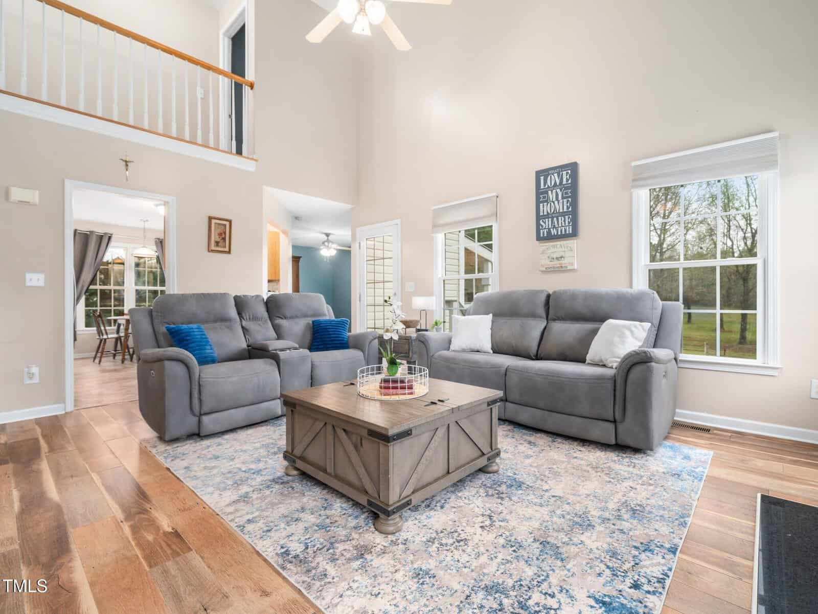A living room with gray sofas and blue accent pillows surrounding a wooden coffee table on a patterned area rug. A sign on the wall reads "Love the Home You Share." Large windows and a ceiling fan provide light. A hallway leads to other rooms.