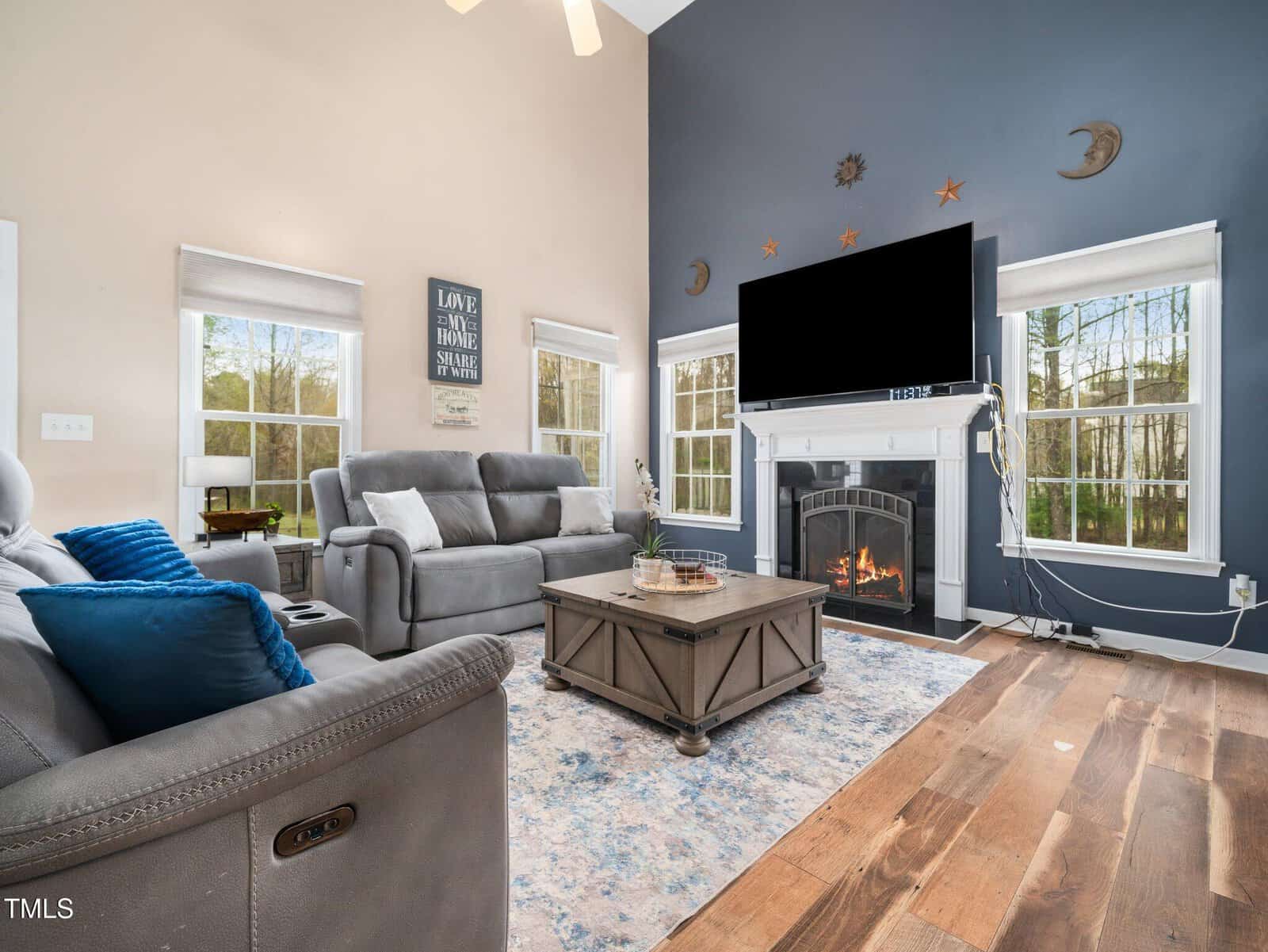 A modern living room with gray sofas, a wooden coffee table, and a large flat-screen TV above a fireplace. The walls are dual-colored with beige and dark blue. Large windows allow natural light, and a patterned rug covers the wooden floor.