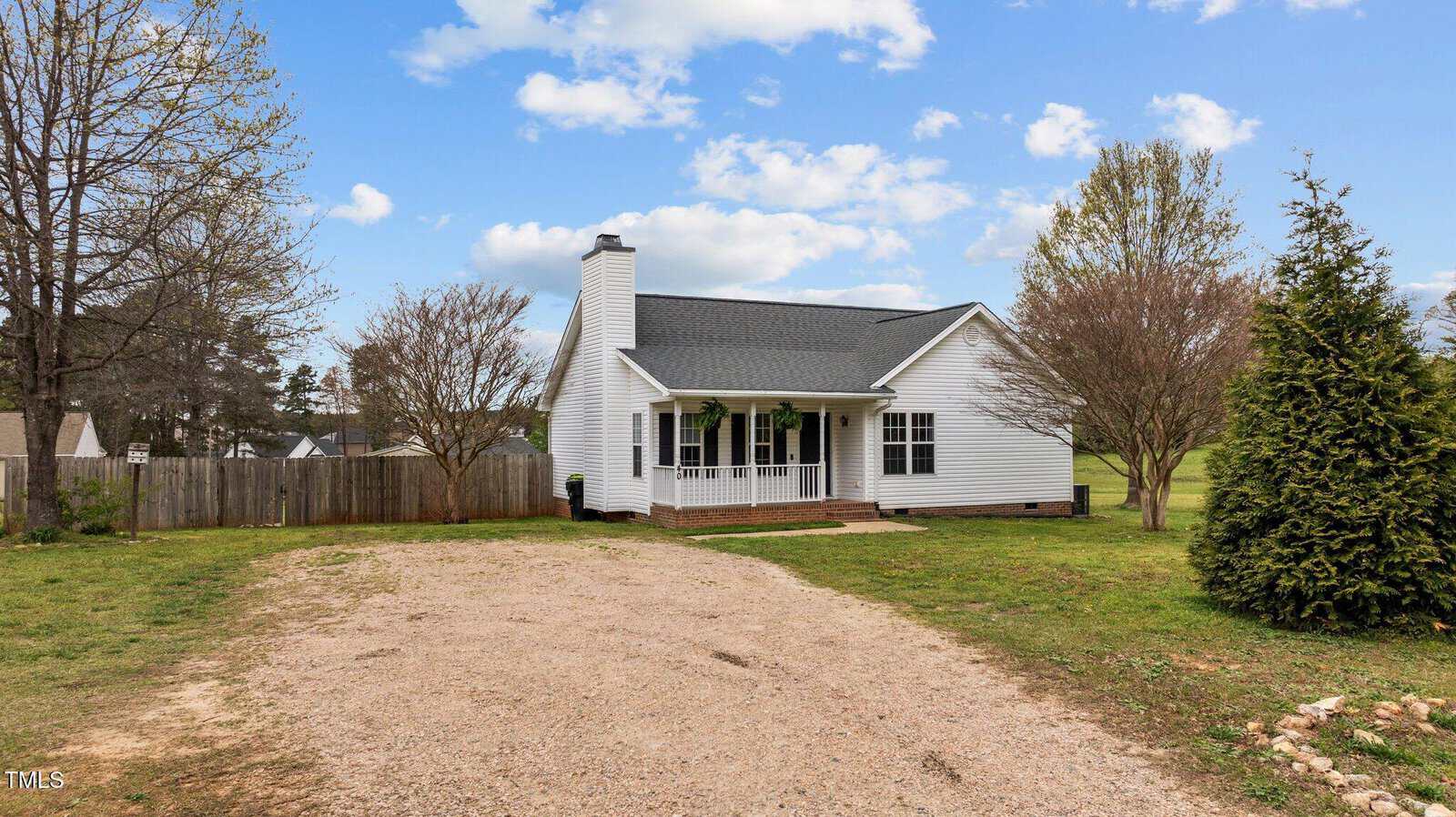 A small white house with a covered front porch, dark roof, and chimney sits at the end of a gravel driveway, surrounded by grass, trees, and a wooden fence under a blue sky with scattered clouds.
