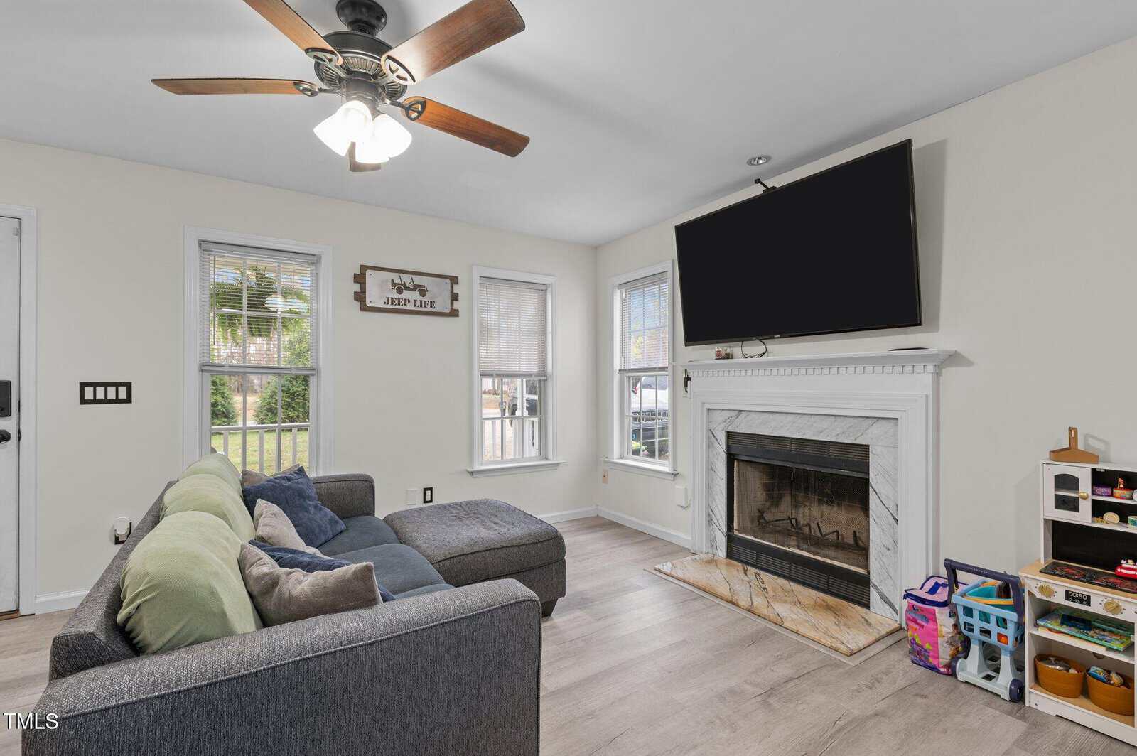 A living room with a sectional sofa, throw pillows, and a ceiling fan. Three windows let in natural light. A TV is mounted above a fireplace. Children's toys and storage bins are in the corner near the mantle.