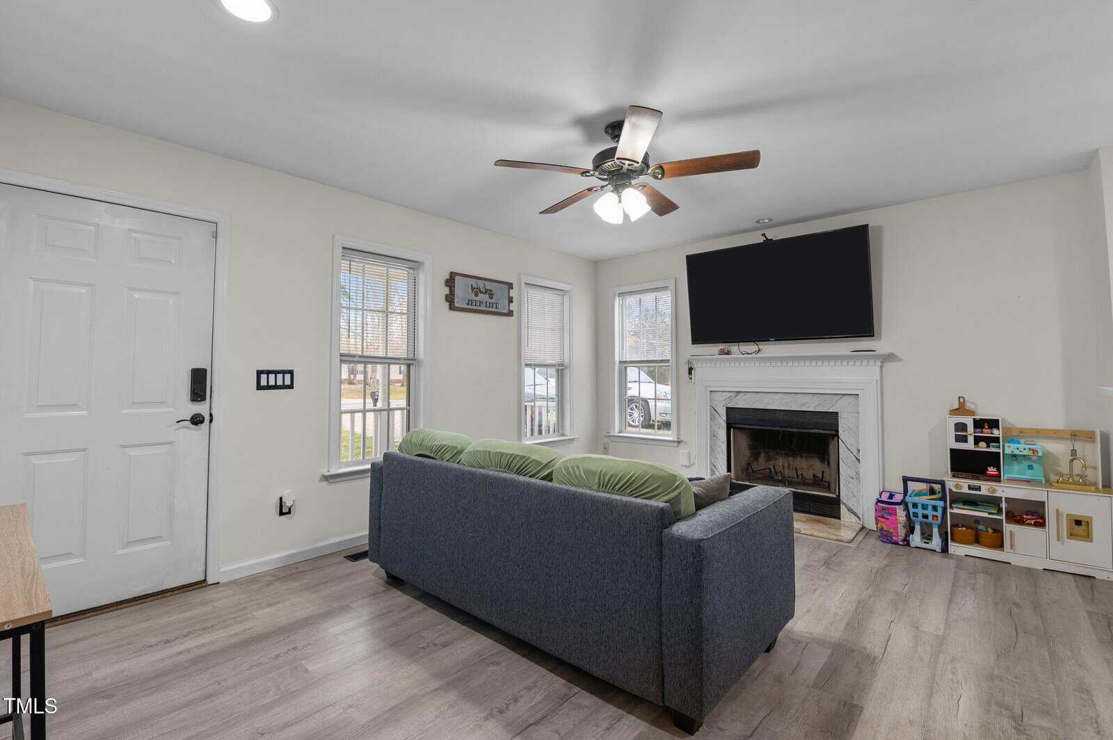 A living room with a gray sofa, a ceiling fan with lights, a fireplace, a wall-mounted TV, toy storage shelves, and three windows letting in natural light. The front door is on the left side of the image.