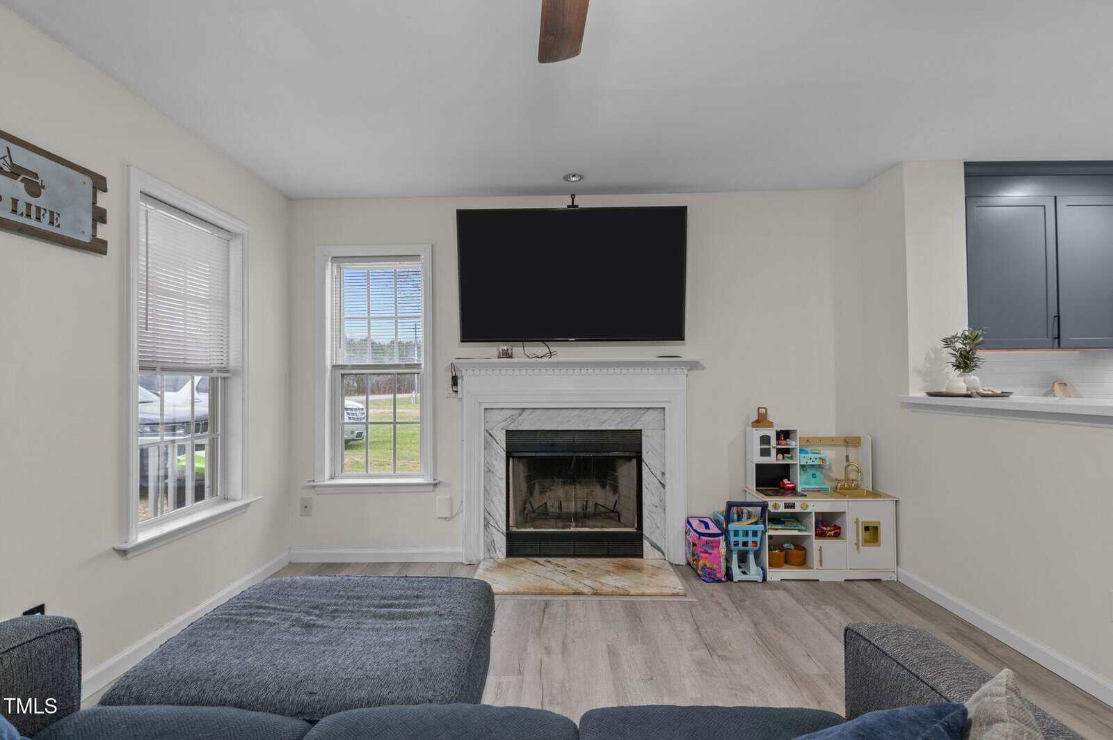 A living room with a gray sectional sofa, a white fireplace with a black TV mounted above, a marble hearth, two windows, light wood flooring, and a children's play kitchen set beside the fireplace.