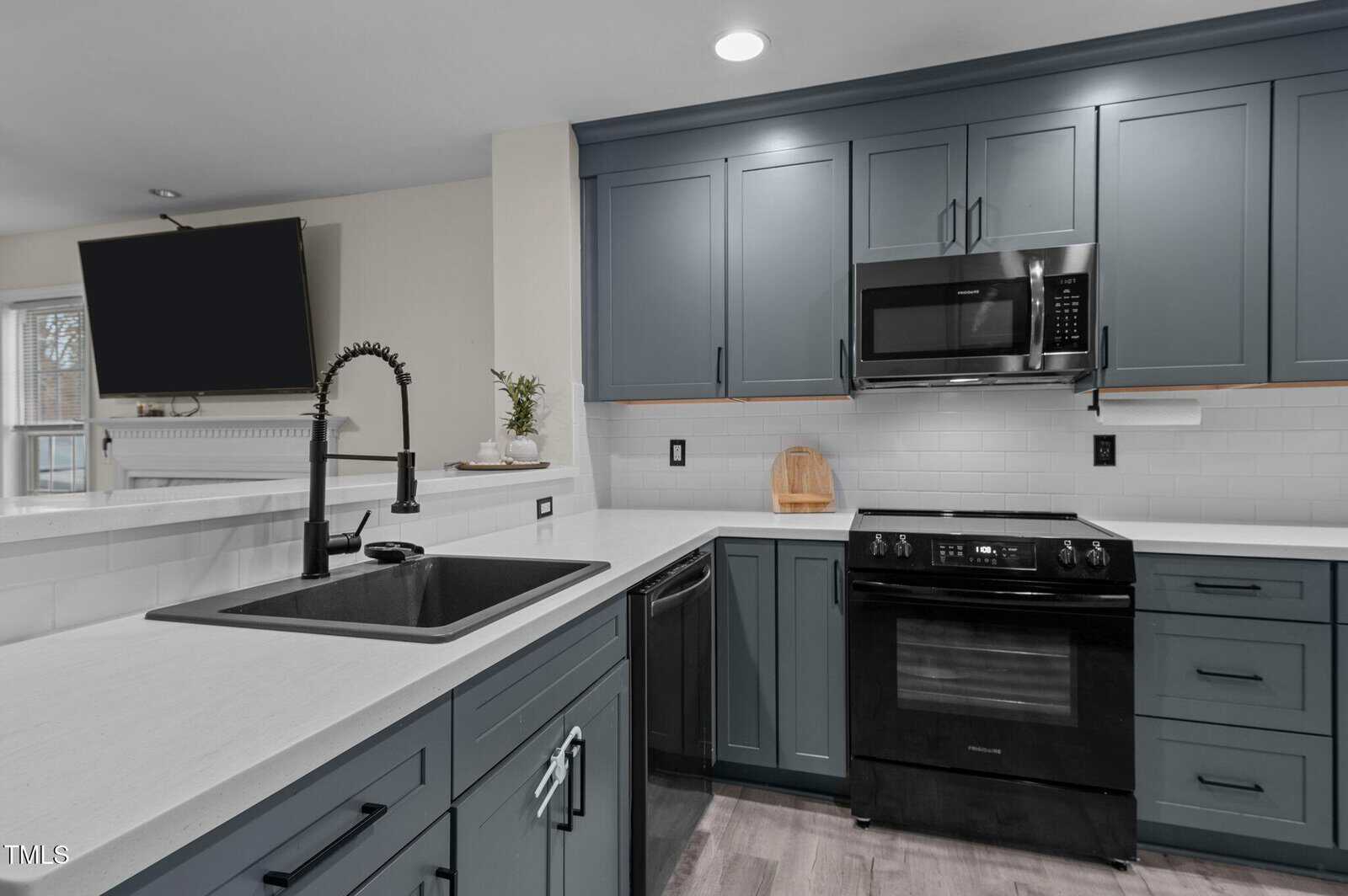 Modern kitchen with blue cabinets, white countertops, black appliances including a stove and microwave, a black sink with a pull-down faucet, a wall-mounted TV, and under-cabinet lighting.