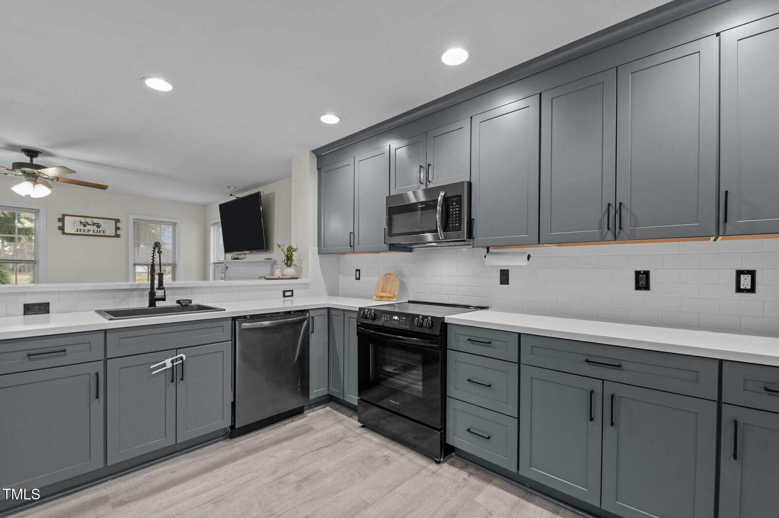 Modern kitchen with gray cabinets, white countertops, stainless steel appliances, white subway tile backsplash, black hardware, and light wood flooring. Ceiling lights, a mounted TV, and a window are visible in the background.
