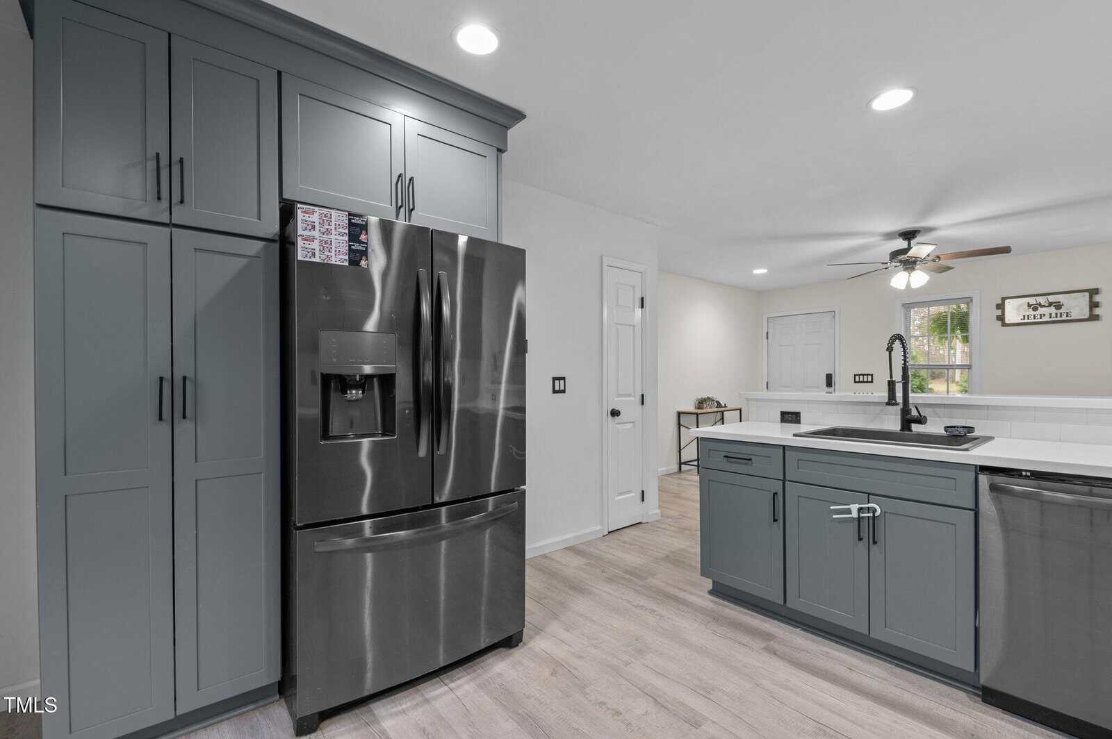 Modern kitchen with gray cabinets, stainless steel refrigerator and dishwasher, light wood flooring, white countertops, and recessed lighting, with a ceiling fan and window in the background.