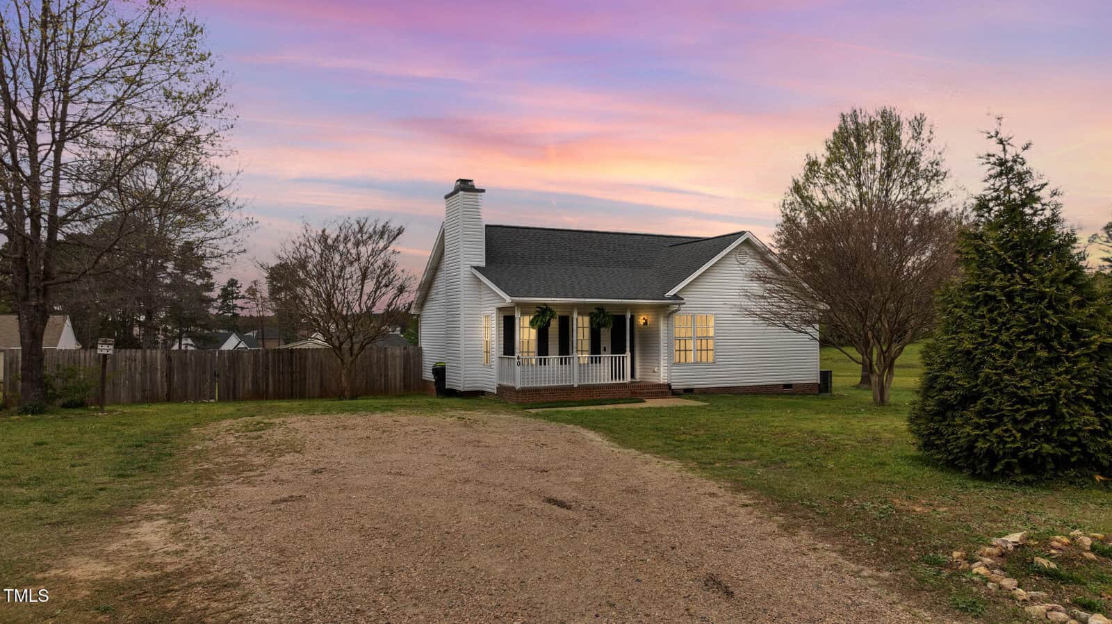 A single-story white house with a front porch sits on a spacious lot with a gravel driveway, surrounded by grass, trees, and a wooden fence at sunset. The sky displays purple, orange, and pink hues.