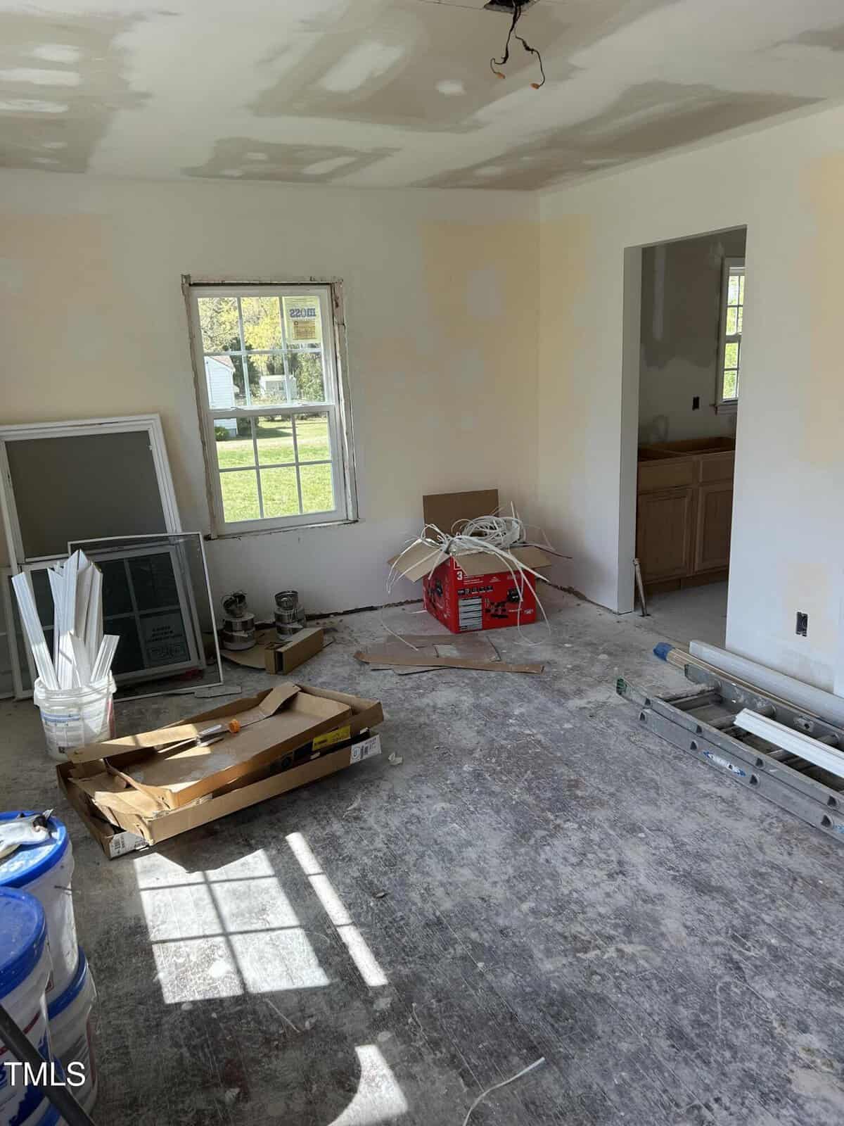 Unfinished room with unpainted walls and a bare floor. Includes a window, various construction materials, tools, a ladder, paint cans, and a partially assembled frame leaning against the wall. Natural light enters through the window.
