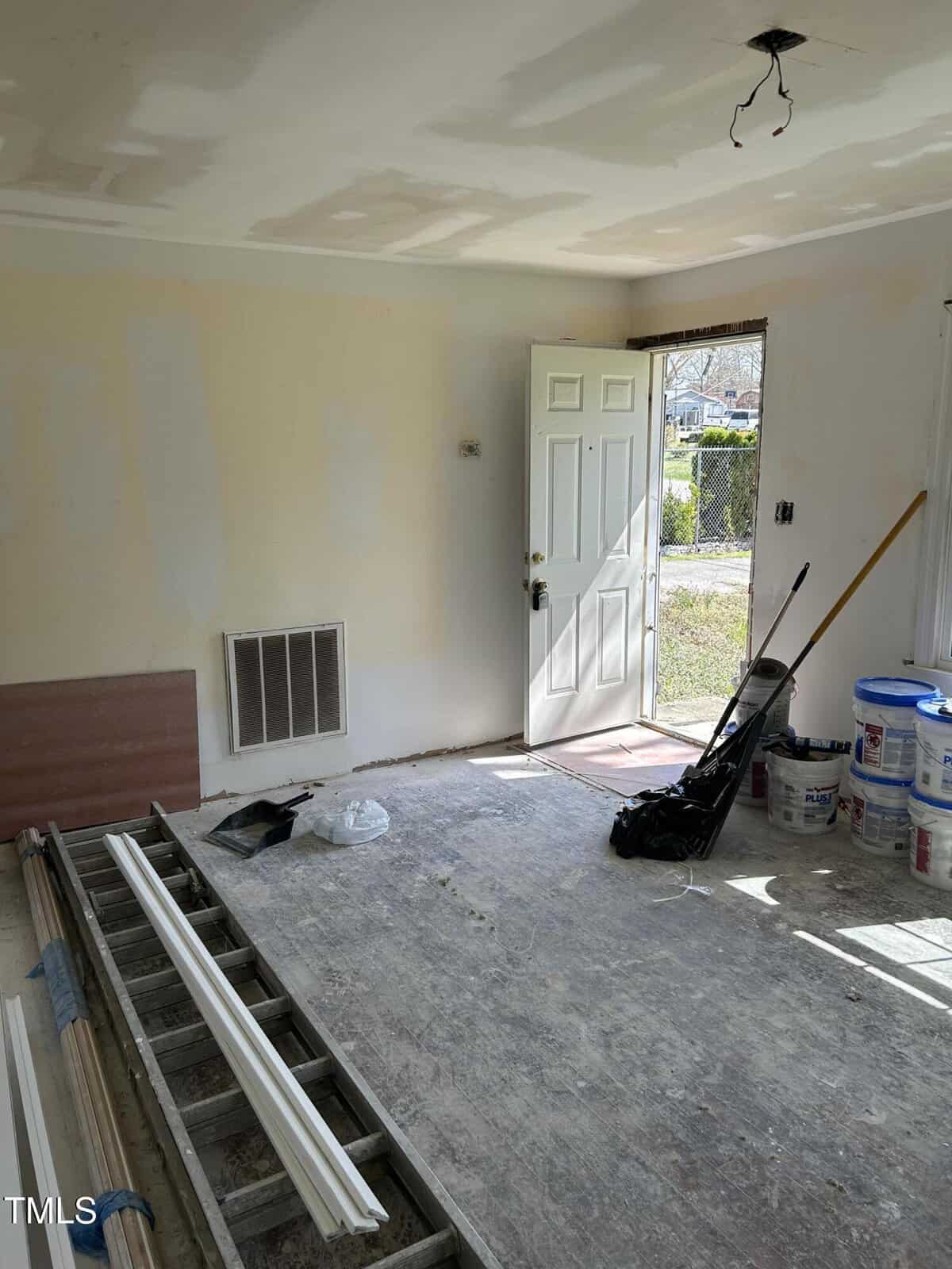 A room under construction with an open door leading outside. The floor is bare, and there are multiple construction materials like planks, paint buckets, and tools scattered around. An electrical fixture hangs from the ceiling without a light installed.