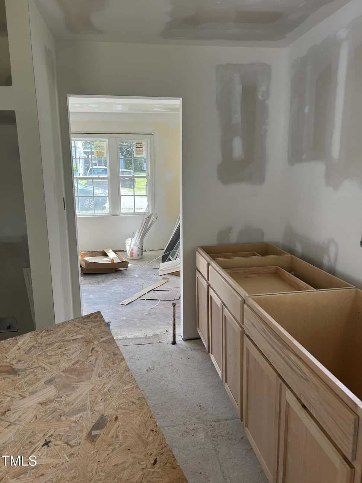 View of a partially completed room under renovation with visible drywall patches. Wooden cabinets are installed on the right, with unfinished countertop space above. Construction materials and tools are visible in adjacent room through the doorway.