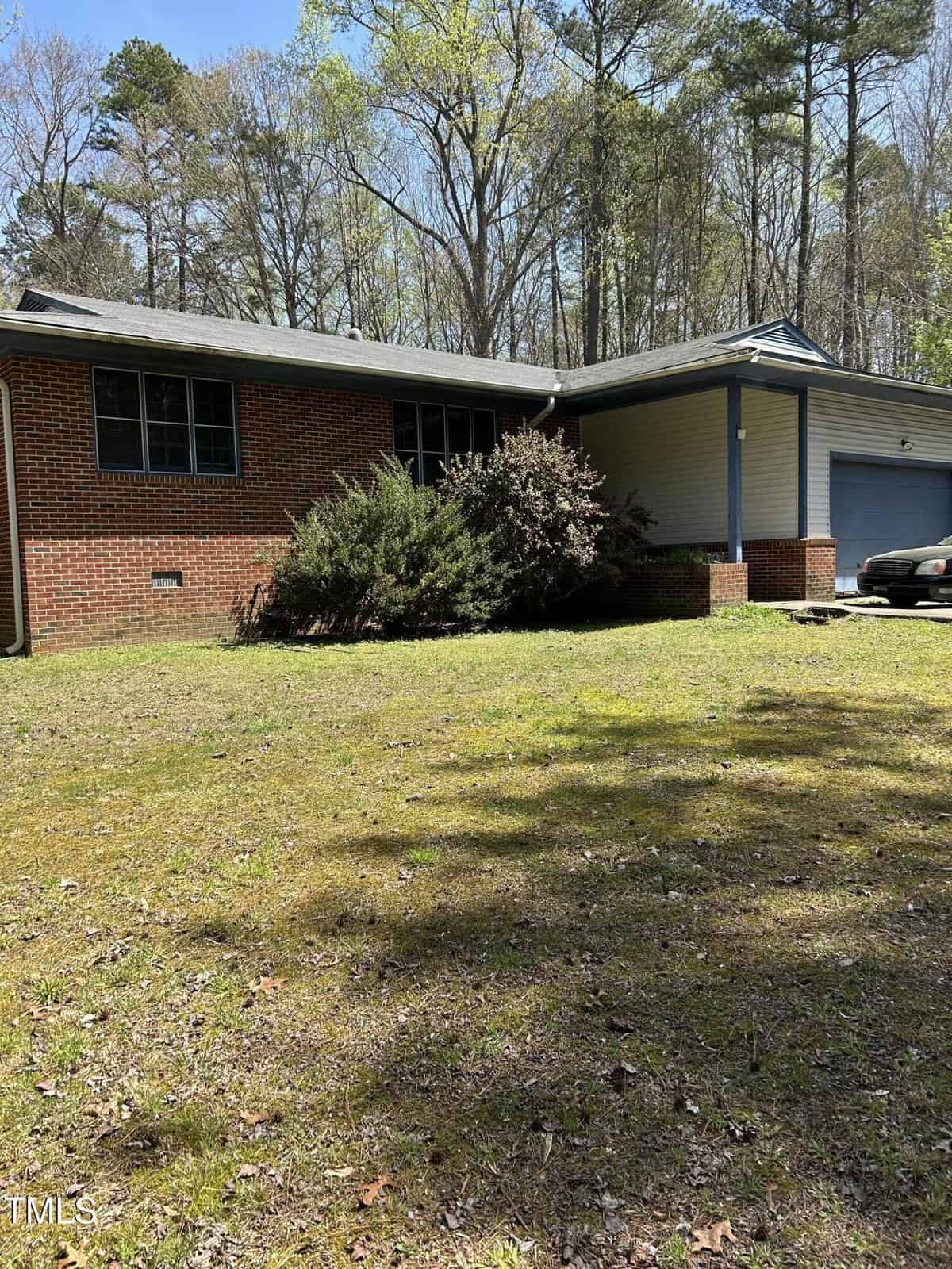 A single-story brick house with white siding is partially obscured by shrubs. There is a two-car garage on the right. The front yard has a patchy lawn with trees in the background under a clear sky.