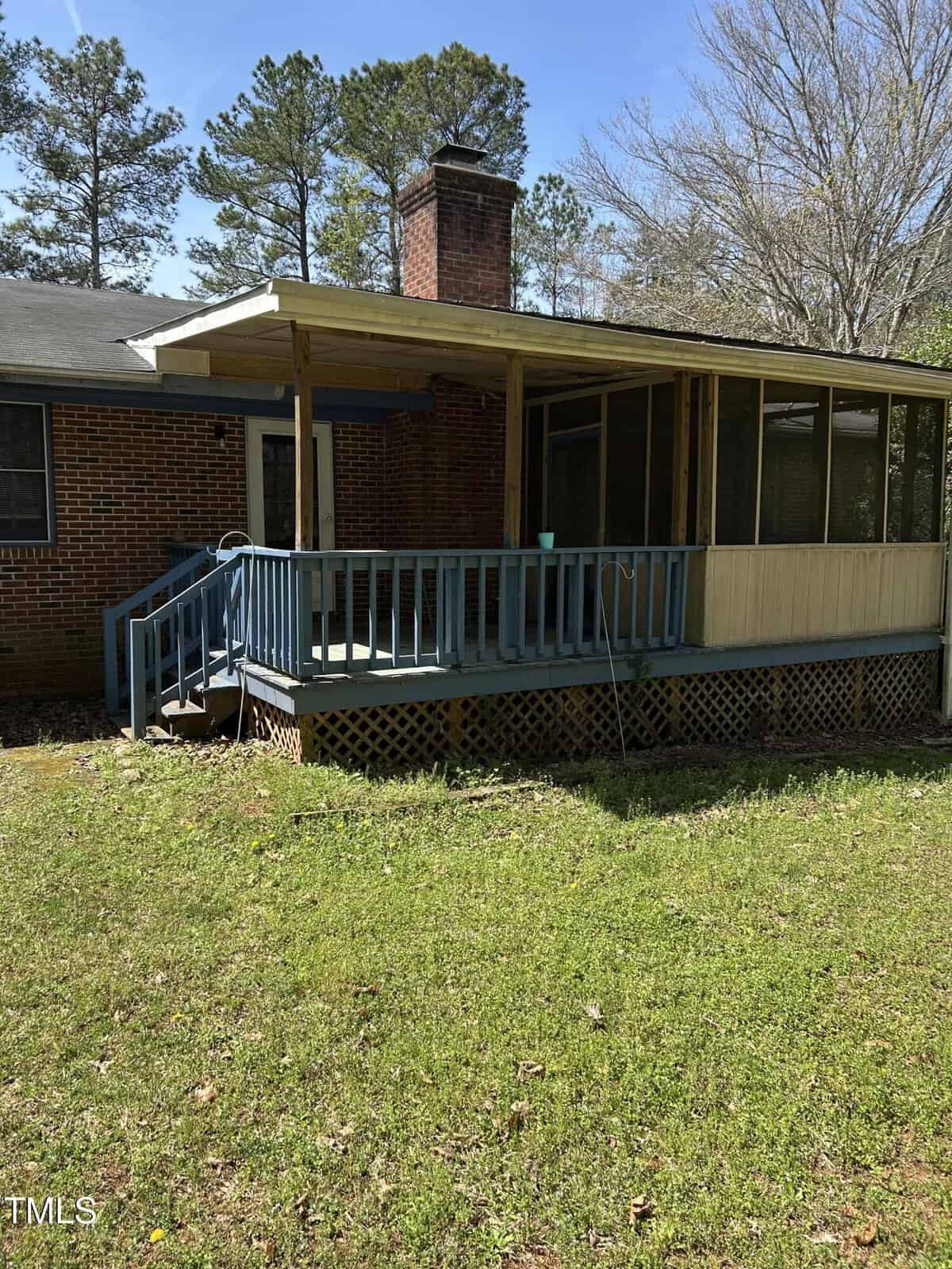 A brick house with a covered porch featuring a blue railing and steps. The porch has a partially wooden enclosure. The ground is grassy with trees in the background, and a brick chimney is visible on the house.