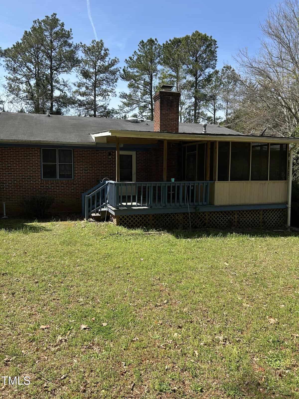 Brick house with a black roof, featuring a screened porch with a chimney and blue railing. The house is surrounded by trees, and the foreground shows a grassy yard. The sky is clear and sunny.