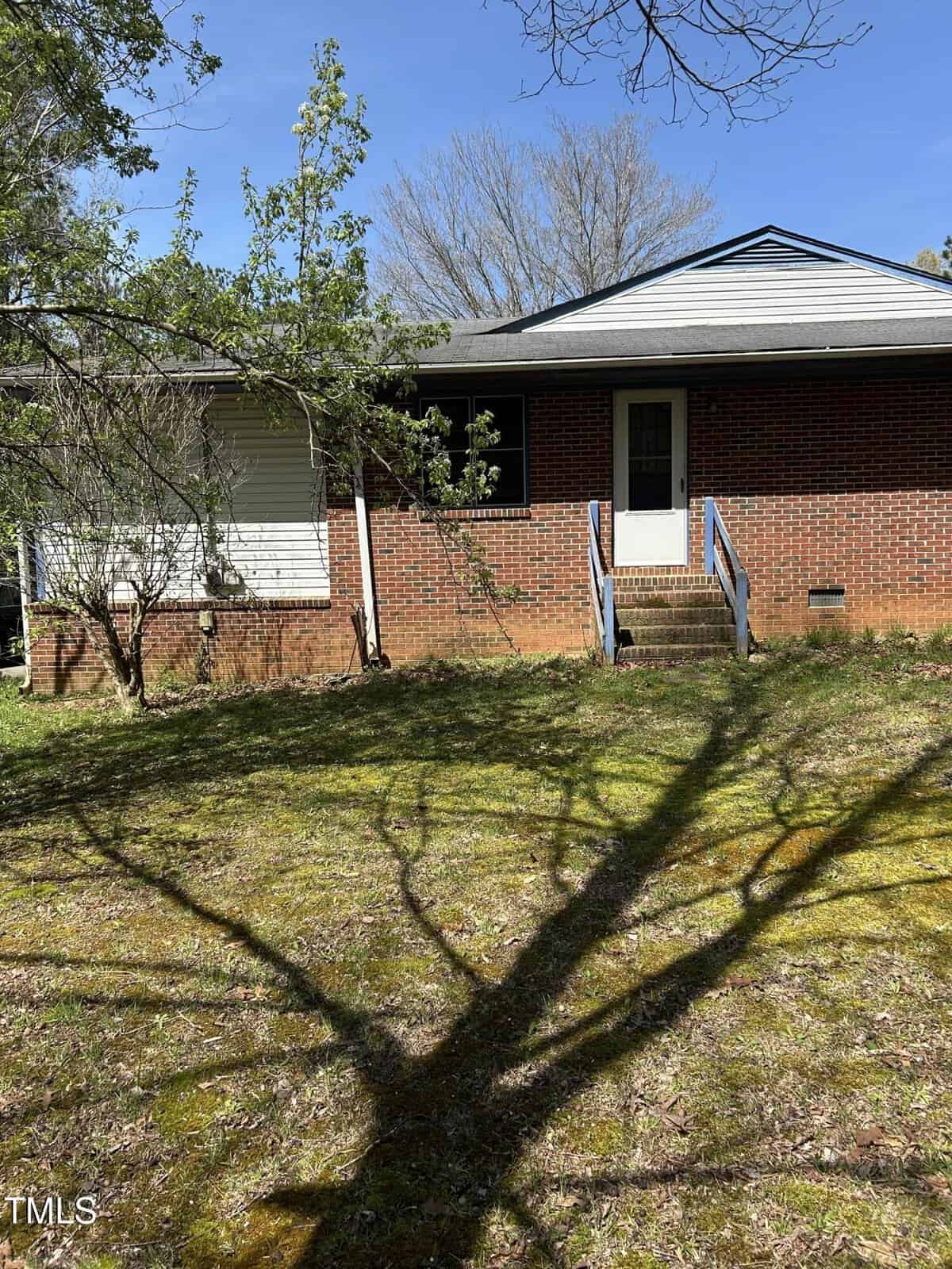 A brick house with a small porch and steps leading up to a white door. There are two windows on the front. A tree in the yard casts a shadow on the grassy area. The sky is clear and blue.