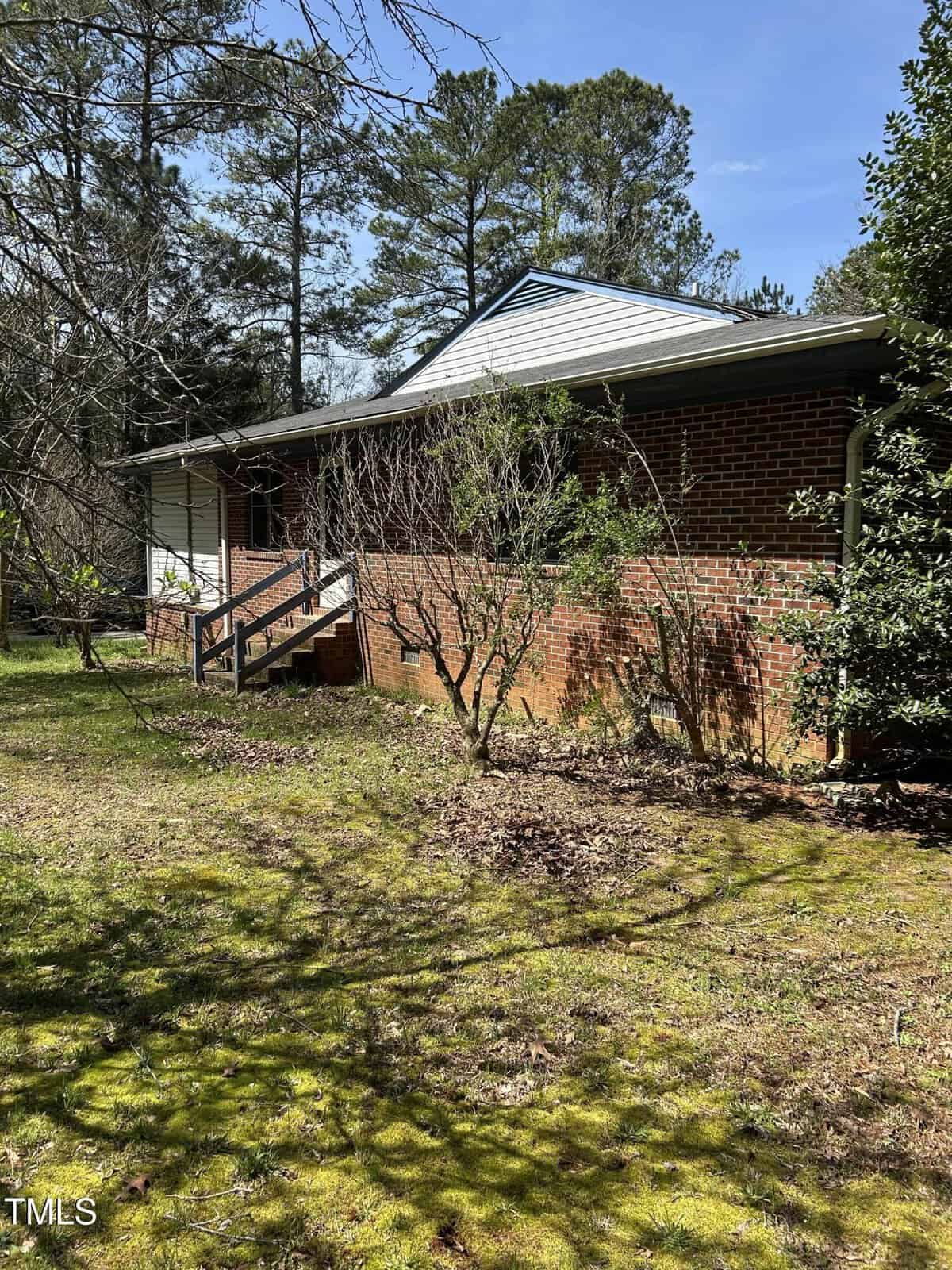 A brick house with a small porch is partially shaded by trees. The house is surrounded by grass and some overgrown shrubs. A staircase with a railing leads up to the porch. The sky is clear and the weather appears sunny.