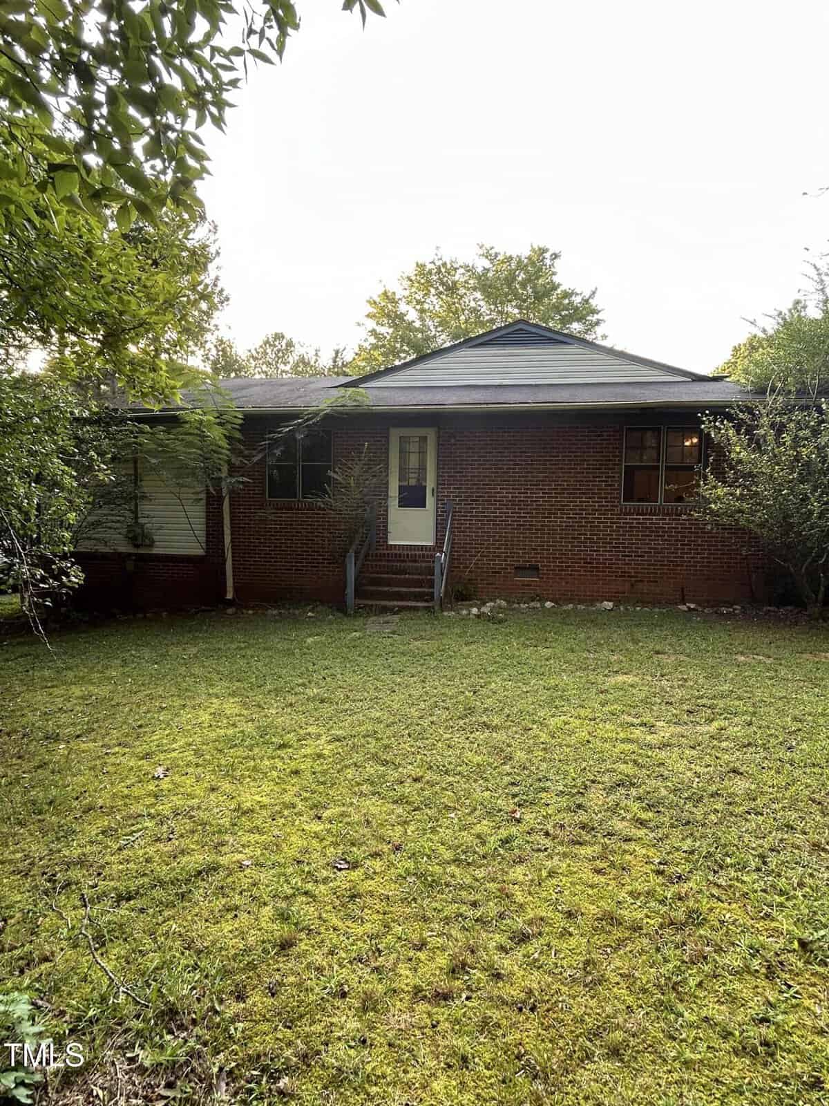A red brick house with a dark roof is partially obscured by trees in the foreground. The house features a small porch with steps leading to a front door and two main windows. The lawn is grassy and slightly overgrown.