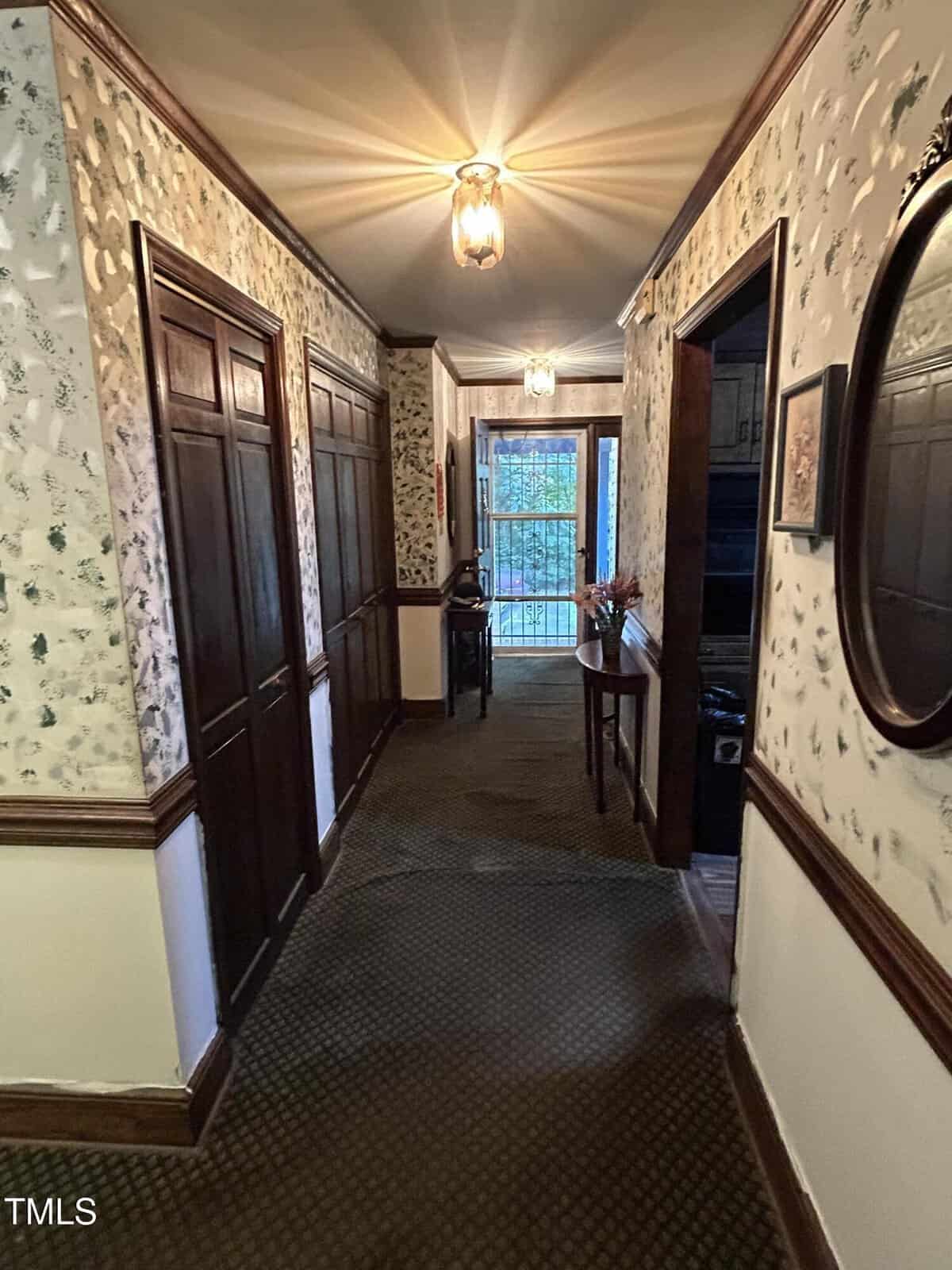 A narrow hallway with patterned wallpaper and brown trim features several closed wooden doors on the left. A table with decorative items is visible ahead, near an open doorway leading to a space with a metal railing and window. A round mirror is on the right wall.
