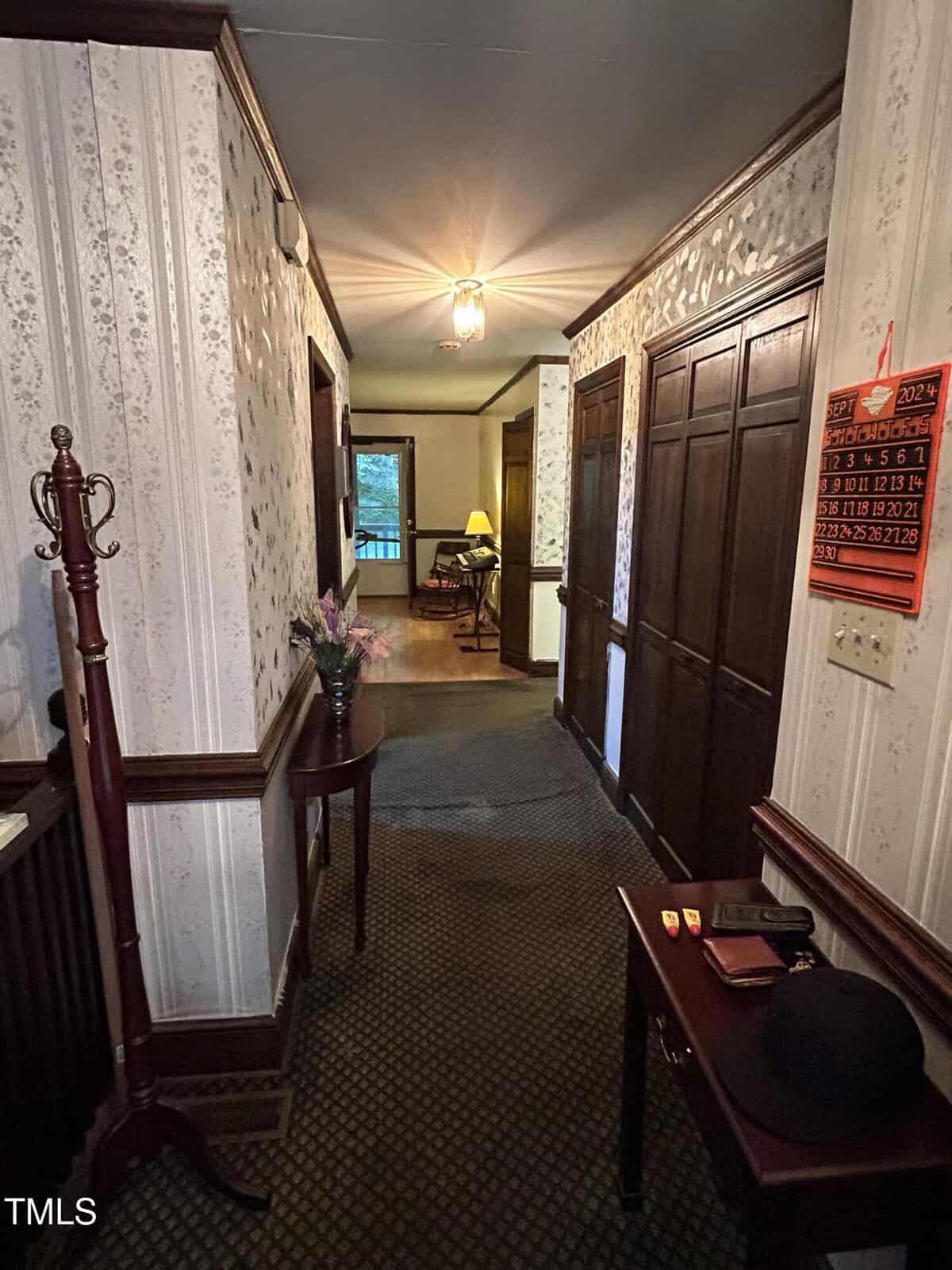 A hallway with floral wallpaper, a light fixture on the ceiling, a coat rack, and a small table. A calendar hangs on a wall. There is a hat and some items on the table. A lit lamp and window are visible in an adjoining room.