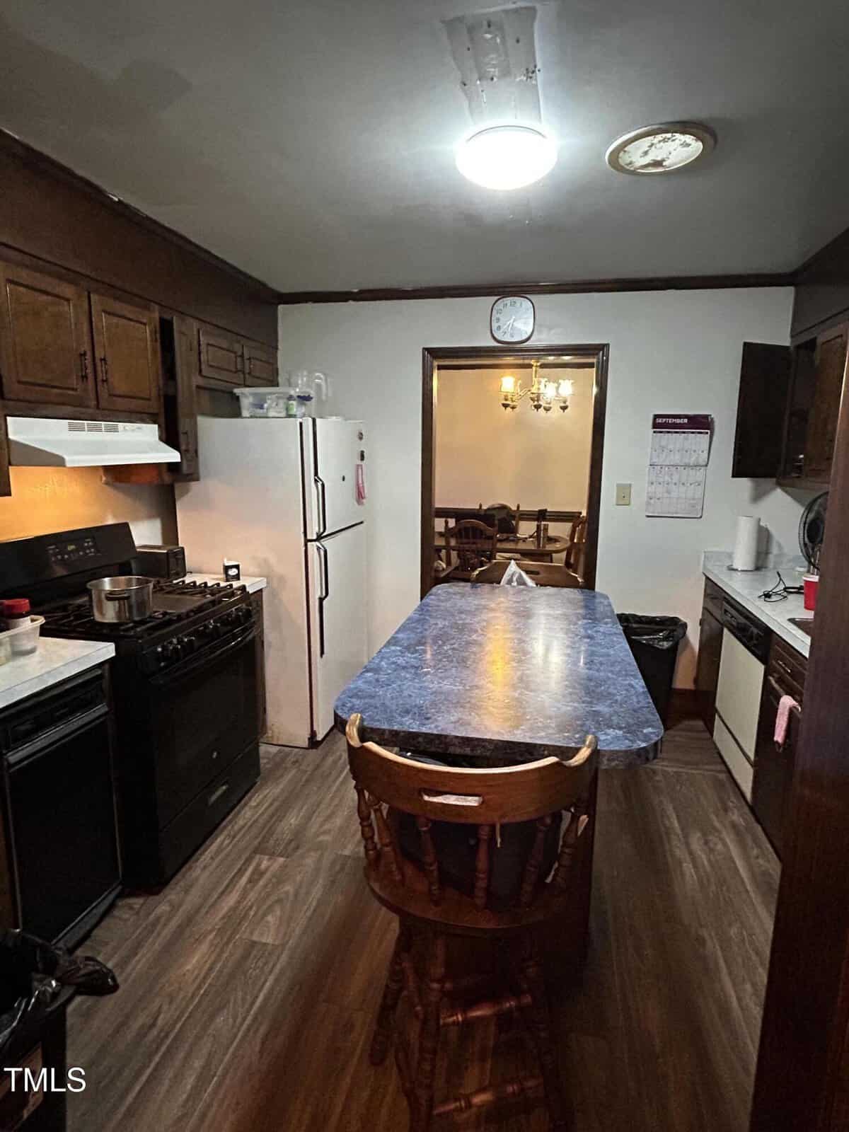 A kitchen with dark wood cabinets, a blue marble-patterned island, a gas stove, white fridge, and dishwasher. Hardwood floors run throughout. A clock and calendar are on the walls. A wooden dining set is visible in the adjacent room.