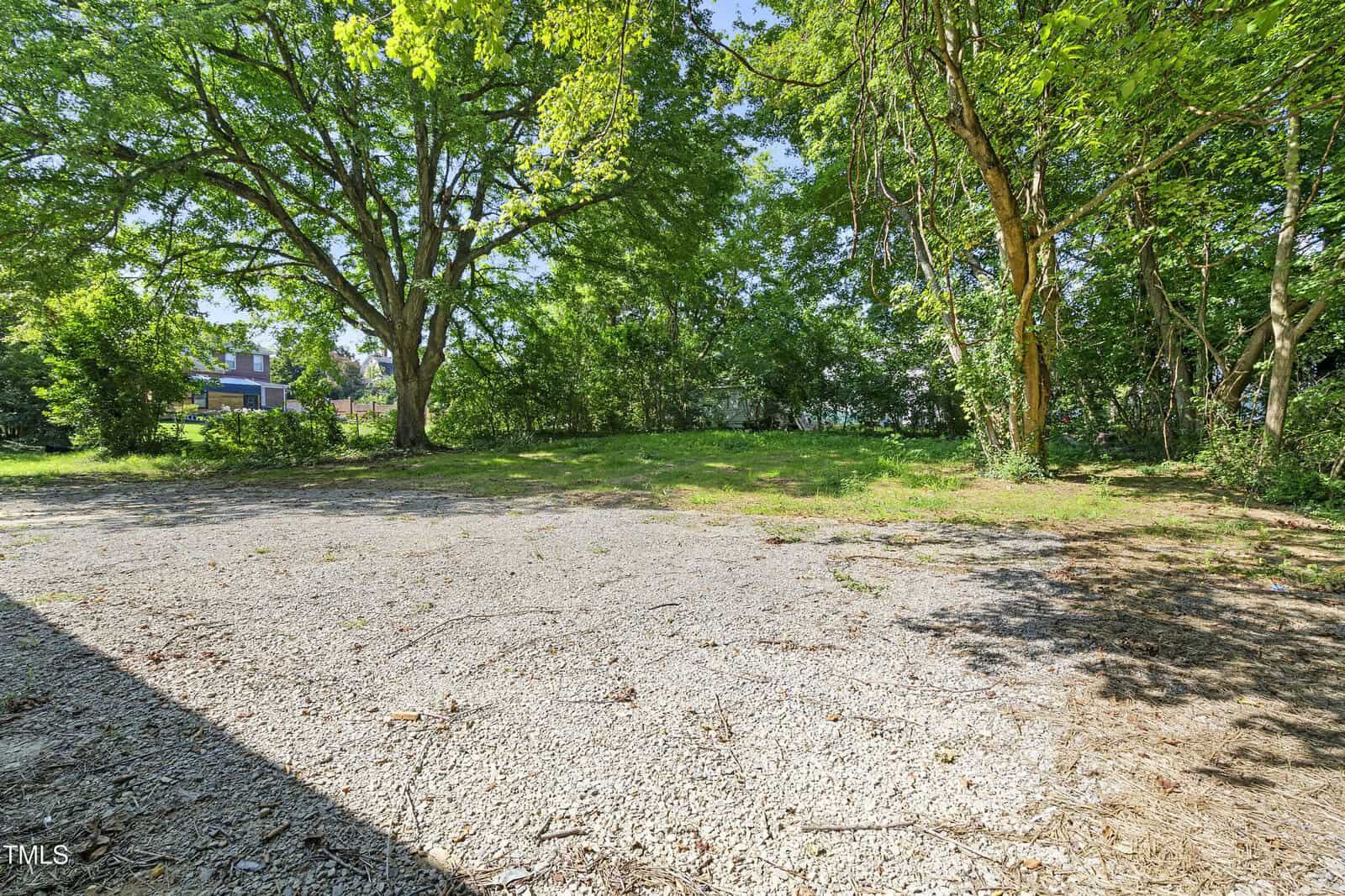 A gravel surface bordered by a grassy area with large trees providing shade defines the serene setting at 129 Burwell Avenue. In the background, part of a house is partially visible among the foliage, while natural sunlight illuminates this peaceful spot in Henderson.