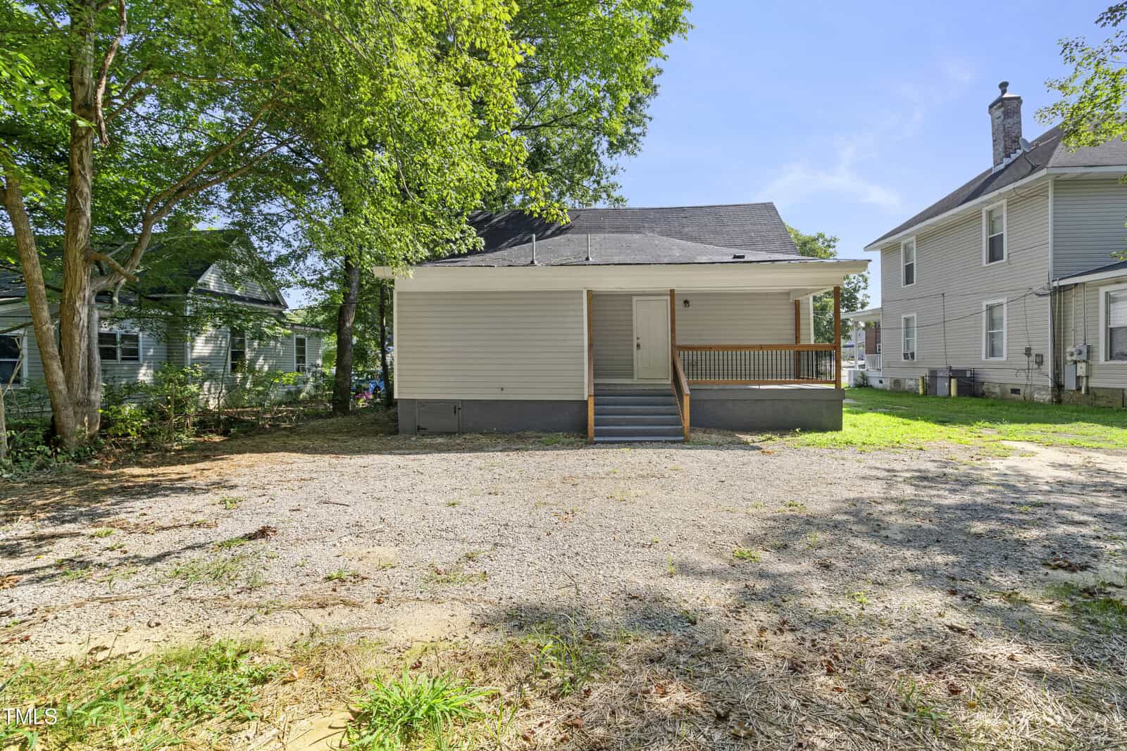 A small, single-story house with gray siding and a covered entry porch is situated in a yard with sparse grass and a gravel area. It is flanked by two other houses and surrounded by a few trees under a clear blue sky.