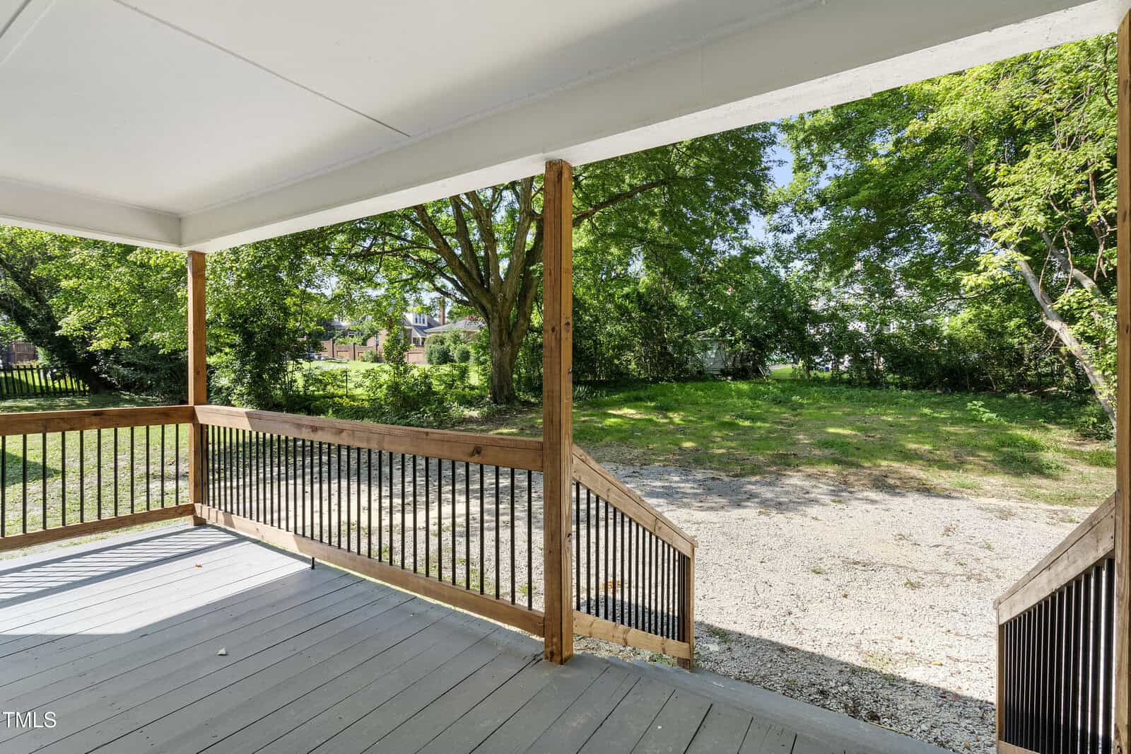 View from a covered porch with wooden railings, overlooking a grassy backyard with trees and a gravel area. The sky is clear and sunny.
