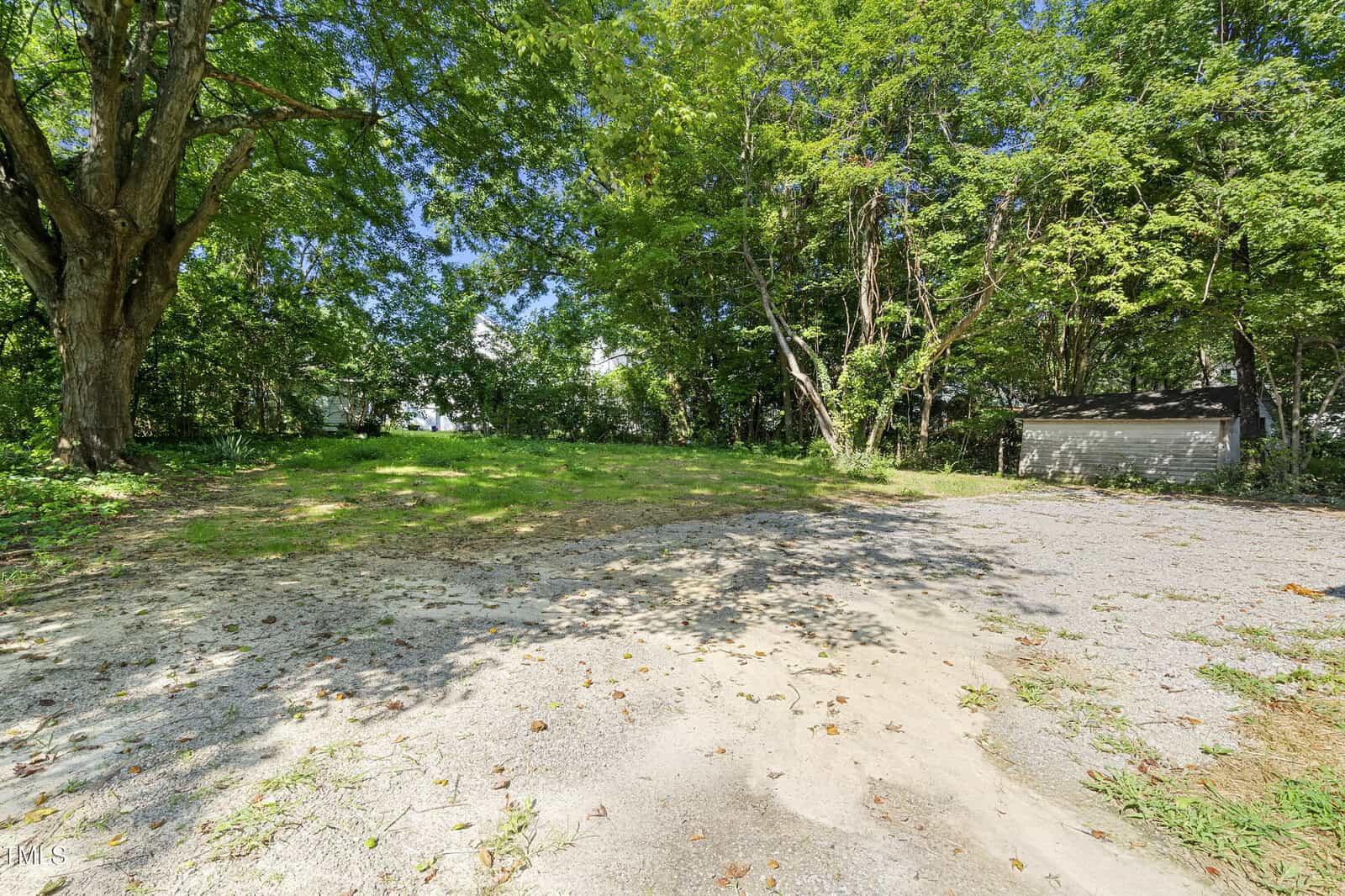 A spacious backyard with green grass and tall trees surrounding the area. The ground is a mix of gravel and dirt in the foreground, leading to the grassy section. A small shed is partially visible on the right side, nestled among the trees.