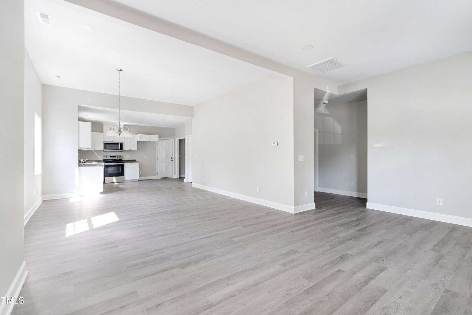Spacious, unfurnished open-concept room with light gray walls and hardwood floors. Kitchen in the background features stainless steel appliances, white cabinets, and an island. Large windows let in natural light.
